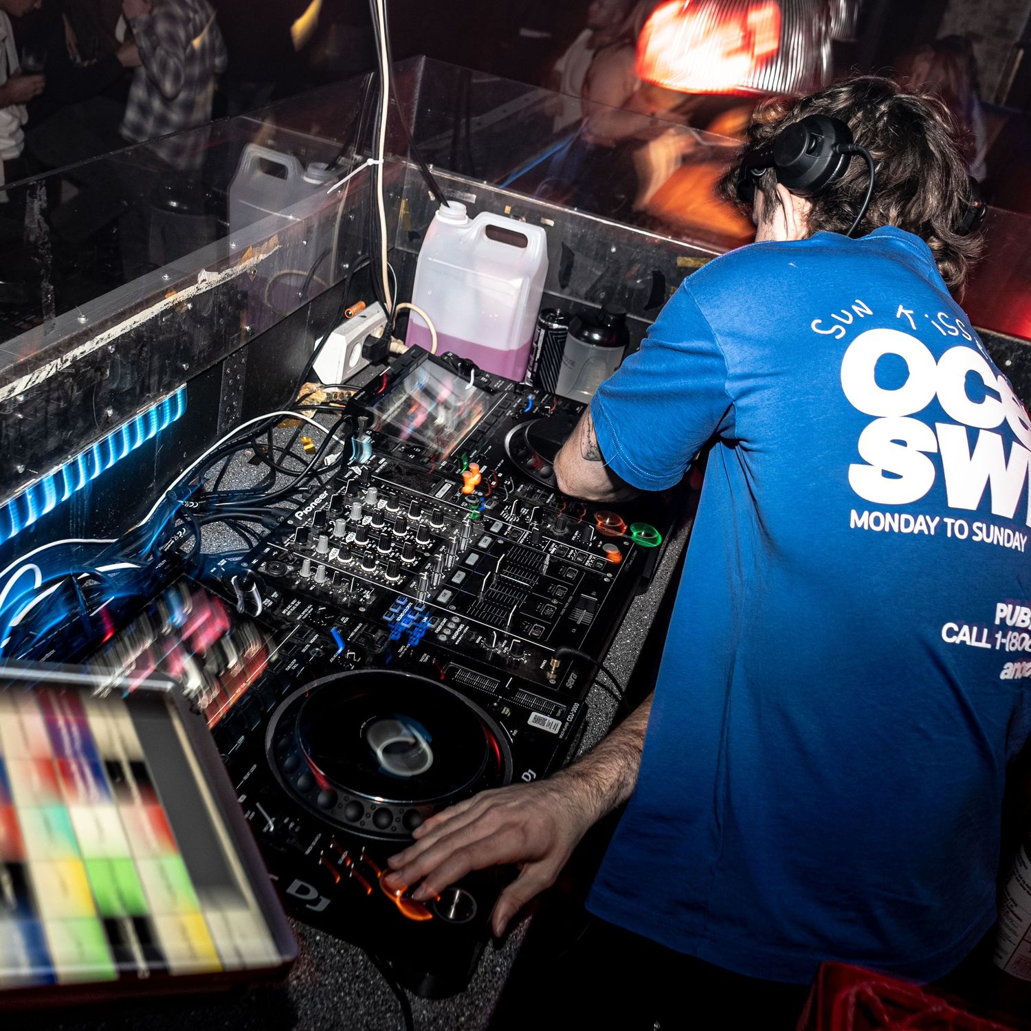 DJ in a blue t-shirt and headphones mixing tracks on a professional DJ controller in a dimly lit nightclub booth.