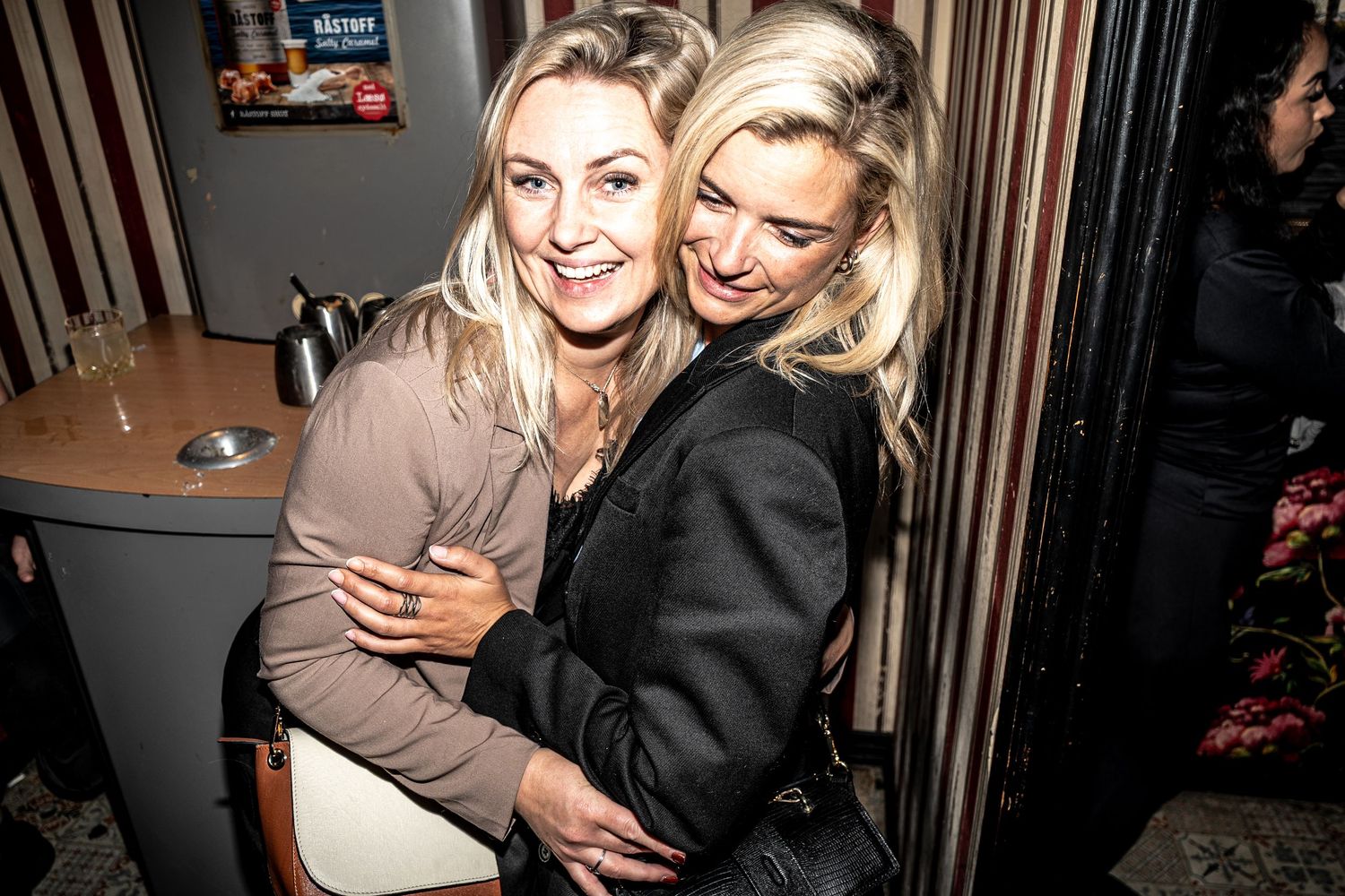 Two smiling blonde women in dark and tan jackets embrace warmly in a cozy, dimly lit bar with a counter in the background.