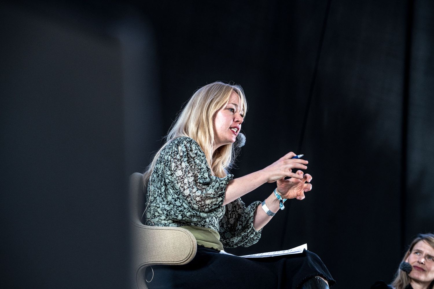 Blonde woman in floral top speaking expressively while seated at an outdoor panel discussion.