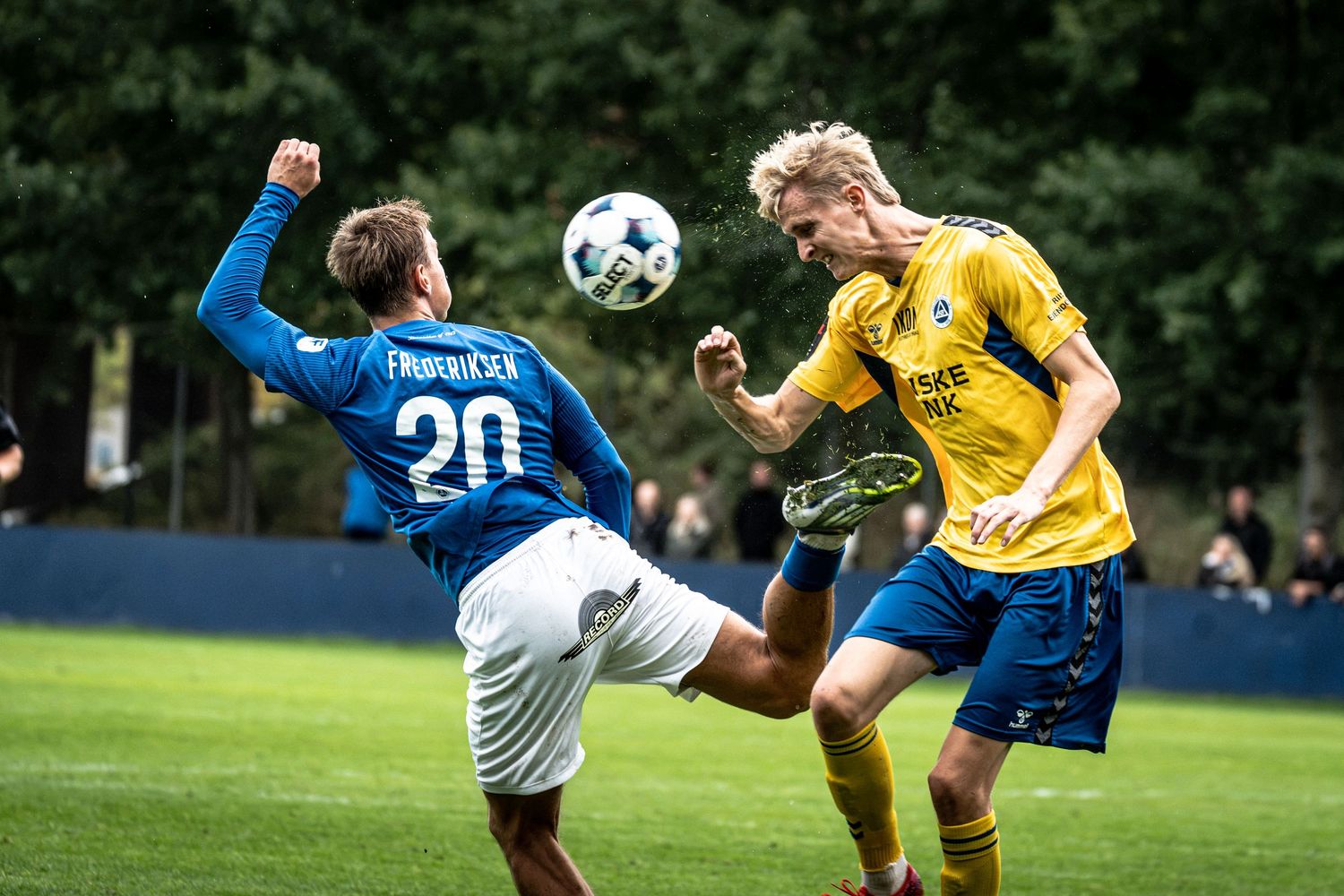 Two soccer players compete for a header, one in blue jersey number 20, the other in yellow, during an outdoor match.