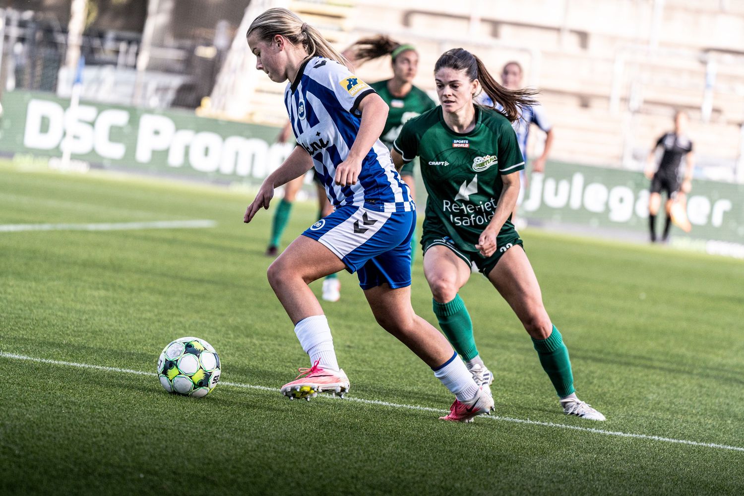 Female soccer player in blue-white kit sprints with ball while green-uniformed opponent gives chase.