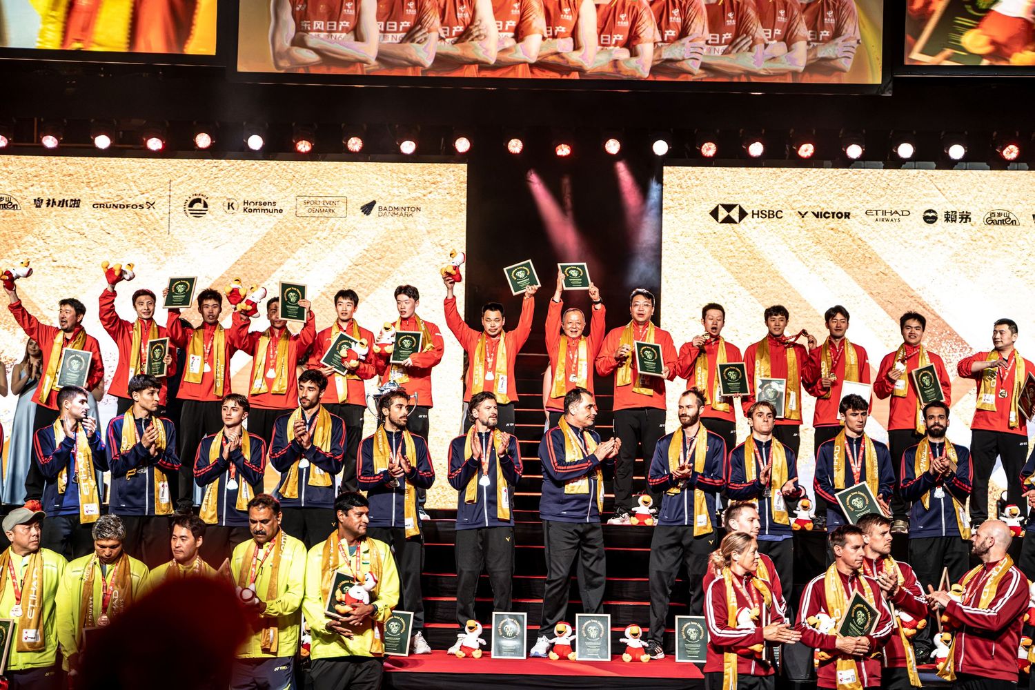 Multiple badminton teams celebrate on stage with medals and awards at championship ceremony.