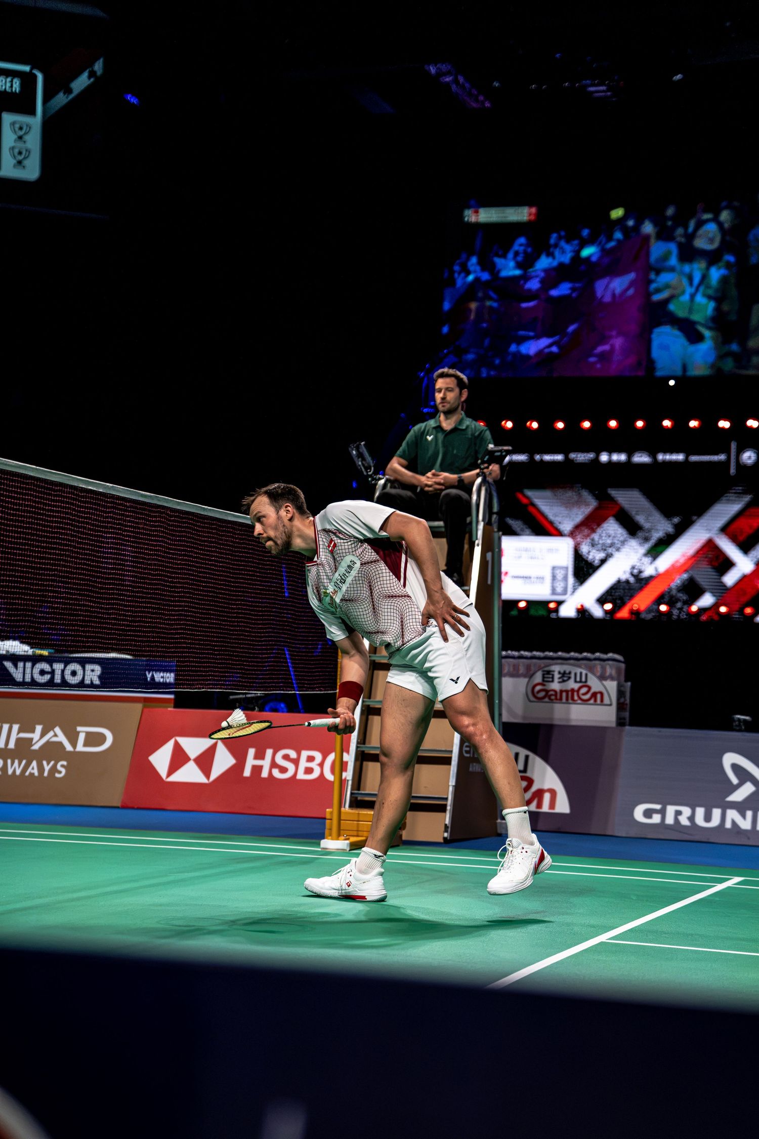 Professional badminton player lunging low to return a shuttlecock during BWF Thomas Cup Finals match in indoor arena.