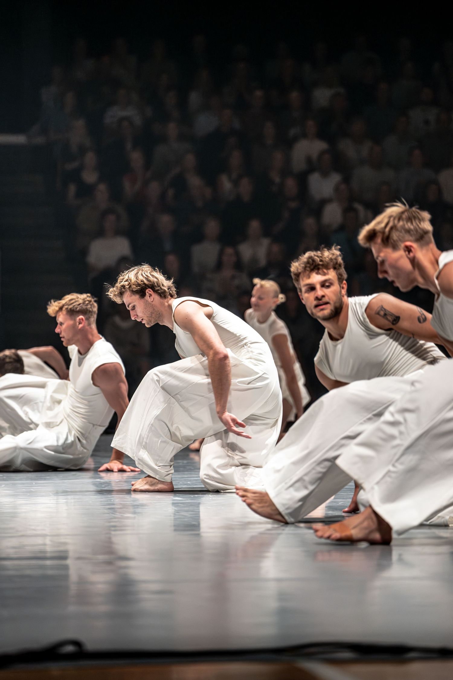 Male contemporary dancers in white costumes crouch on stage floor during group performance.