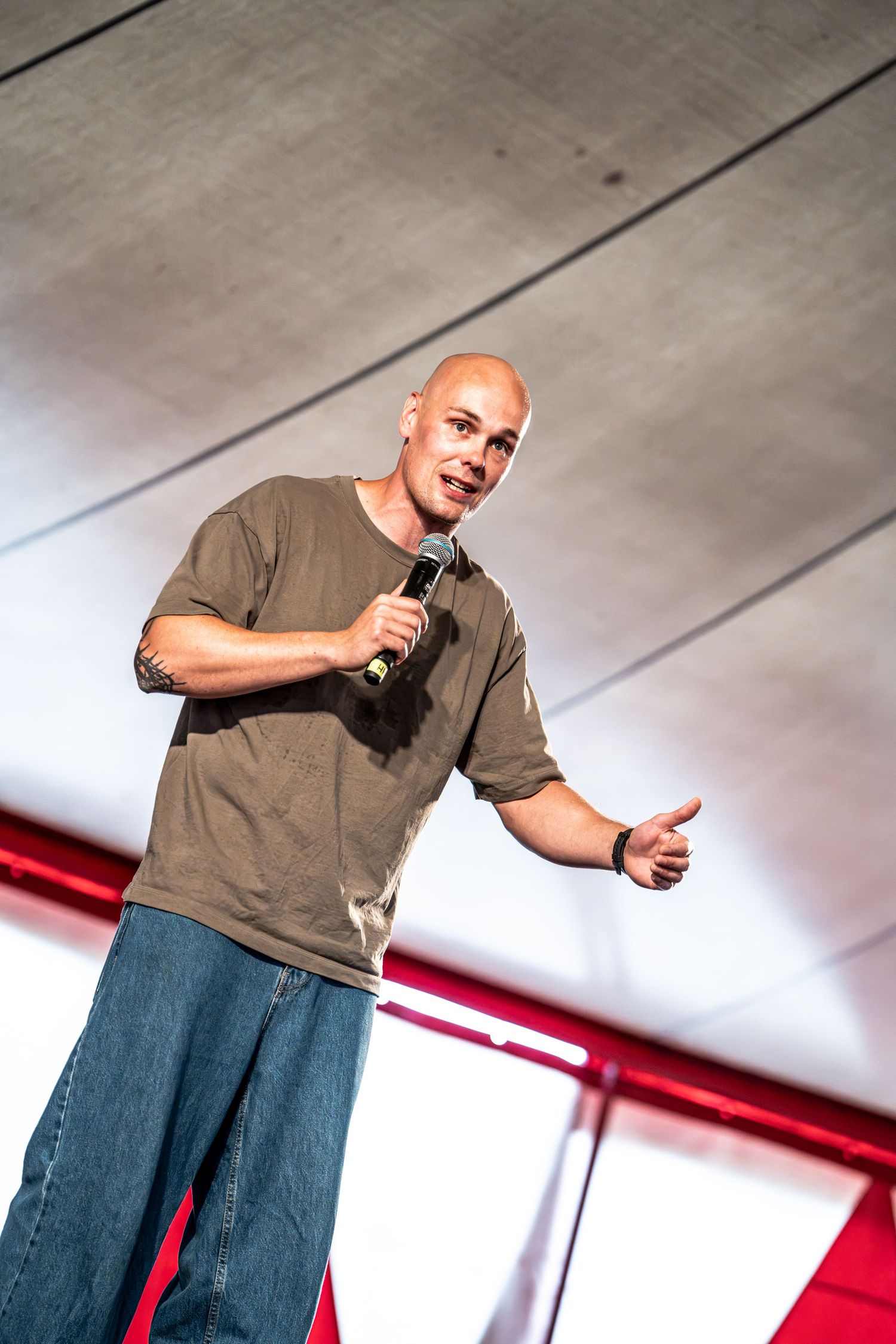 Bald male speaker on stage holding microphone giving thumbs up in olive shirt and jeans.