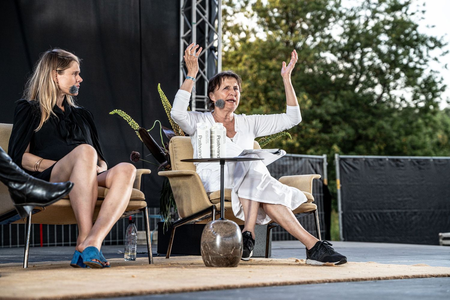 Woman in white raises both hands expressively during an outdoor stage panel discussion.