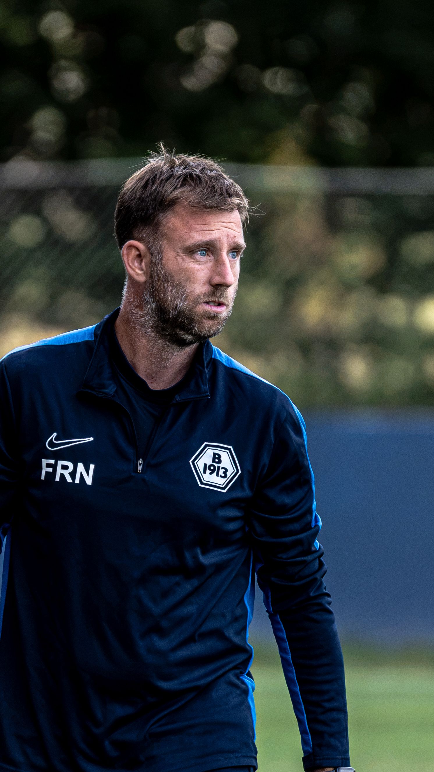Man in navy Nike jacket with B1913 badge stands outdoors on a football pitch looking focused.