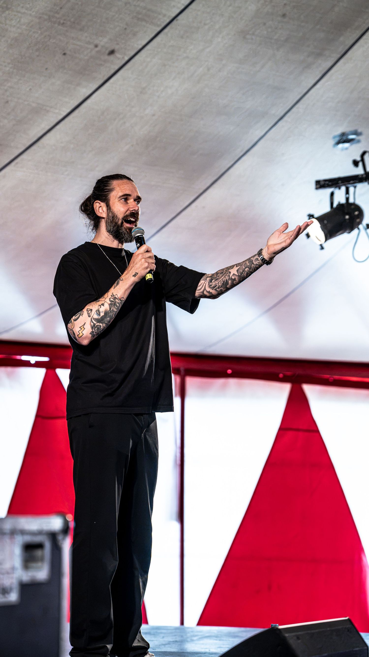 Tattooed bearded performer in black outfit gestures on stage with red triangle backdrop.