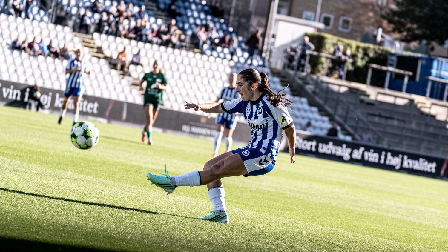 Female footballer in blue and white kit performs powerful kick during sunny outdoor stadium match.