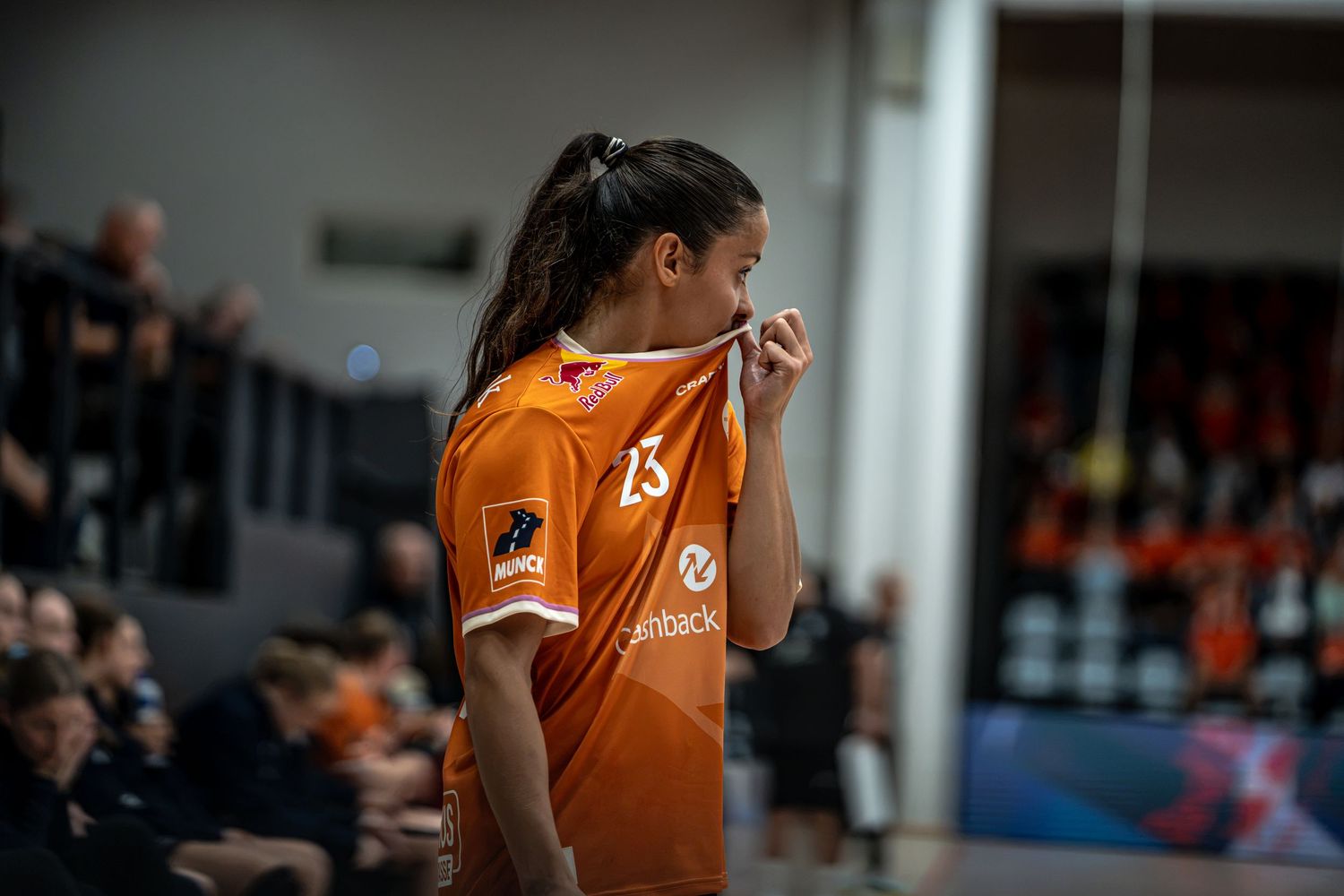 Female handball player in orange jersey biting her shirt collar on the sideline during a match.