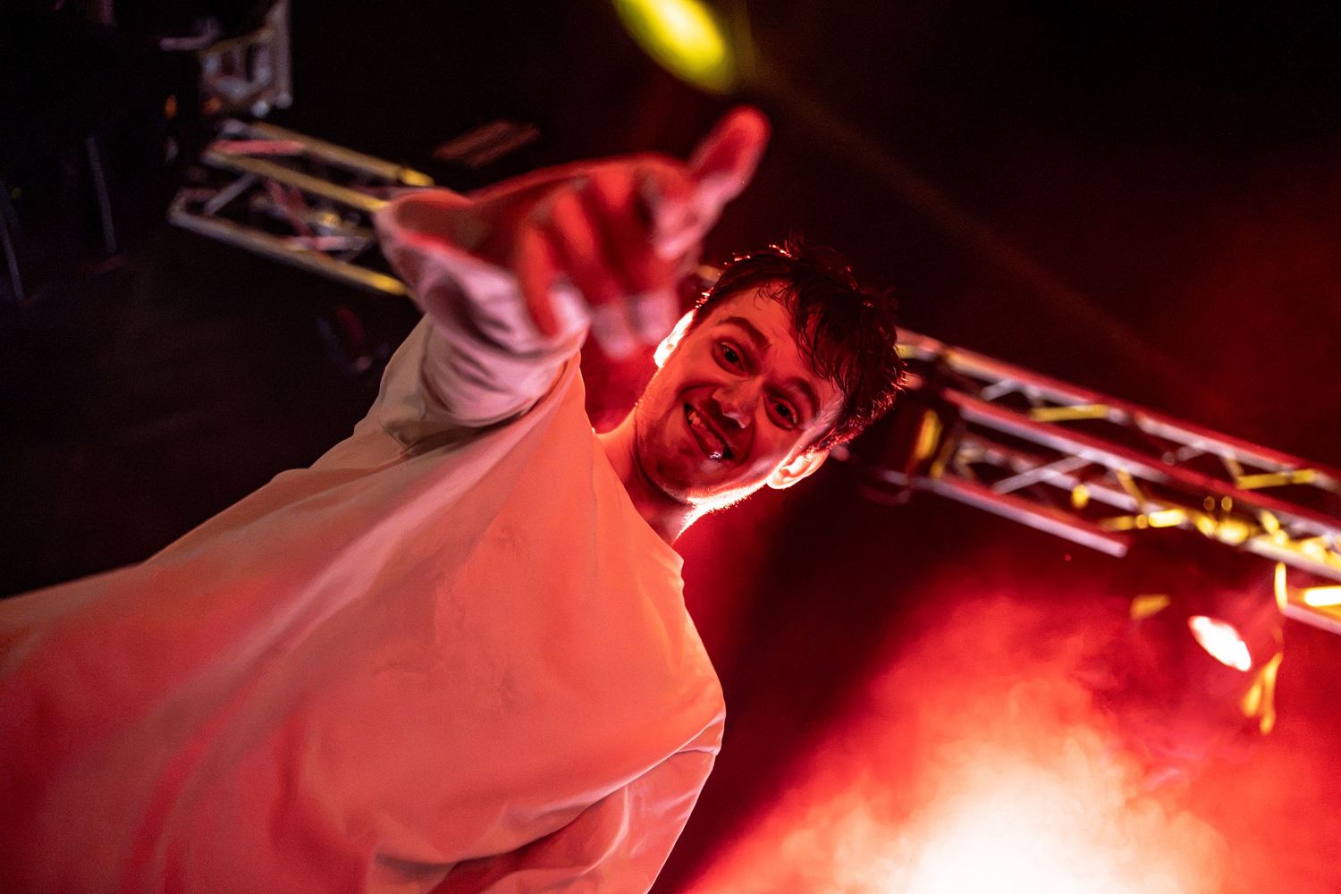 Smiling performer in white outfit reaches toward camera under red stage lighting.