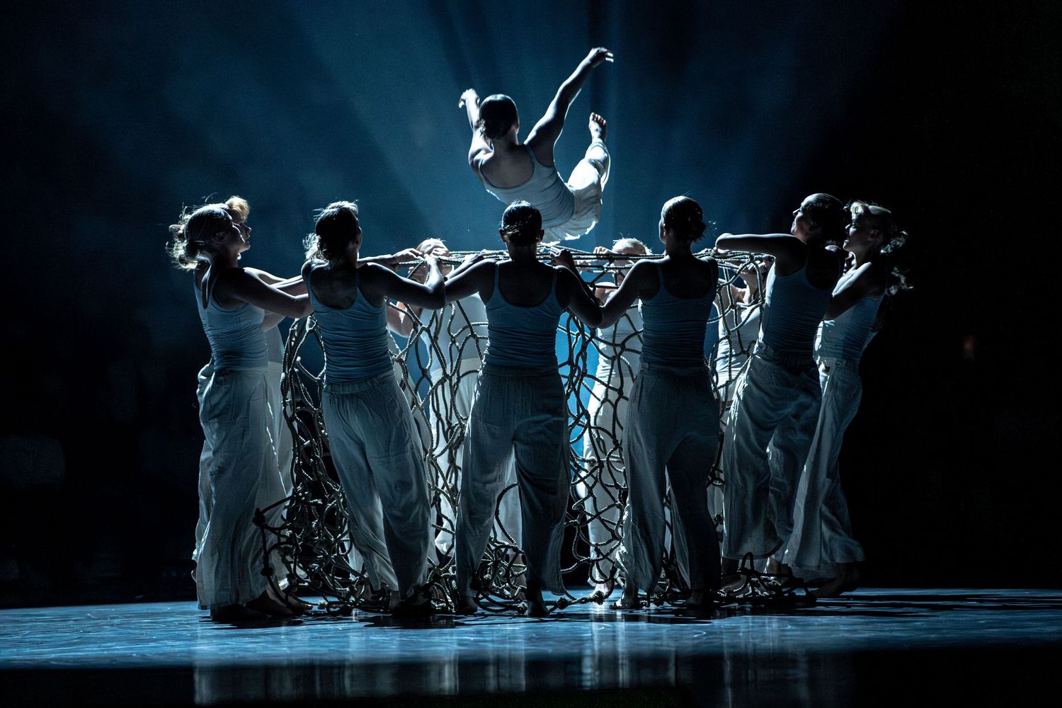 Dance ensemble in white costumes with rope props lifting a performer during a dramatic stage performance.