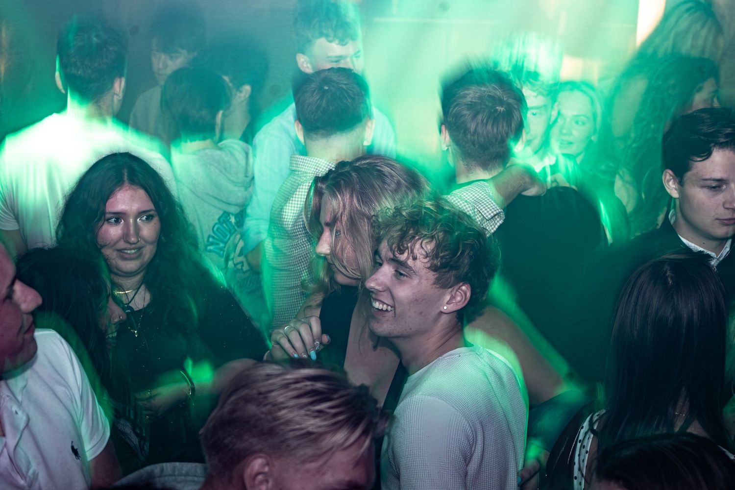 Smiling young people packed together on a dance floor at a lively nightclub party, lit by soft green and warm lights.