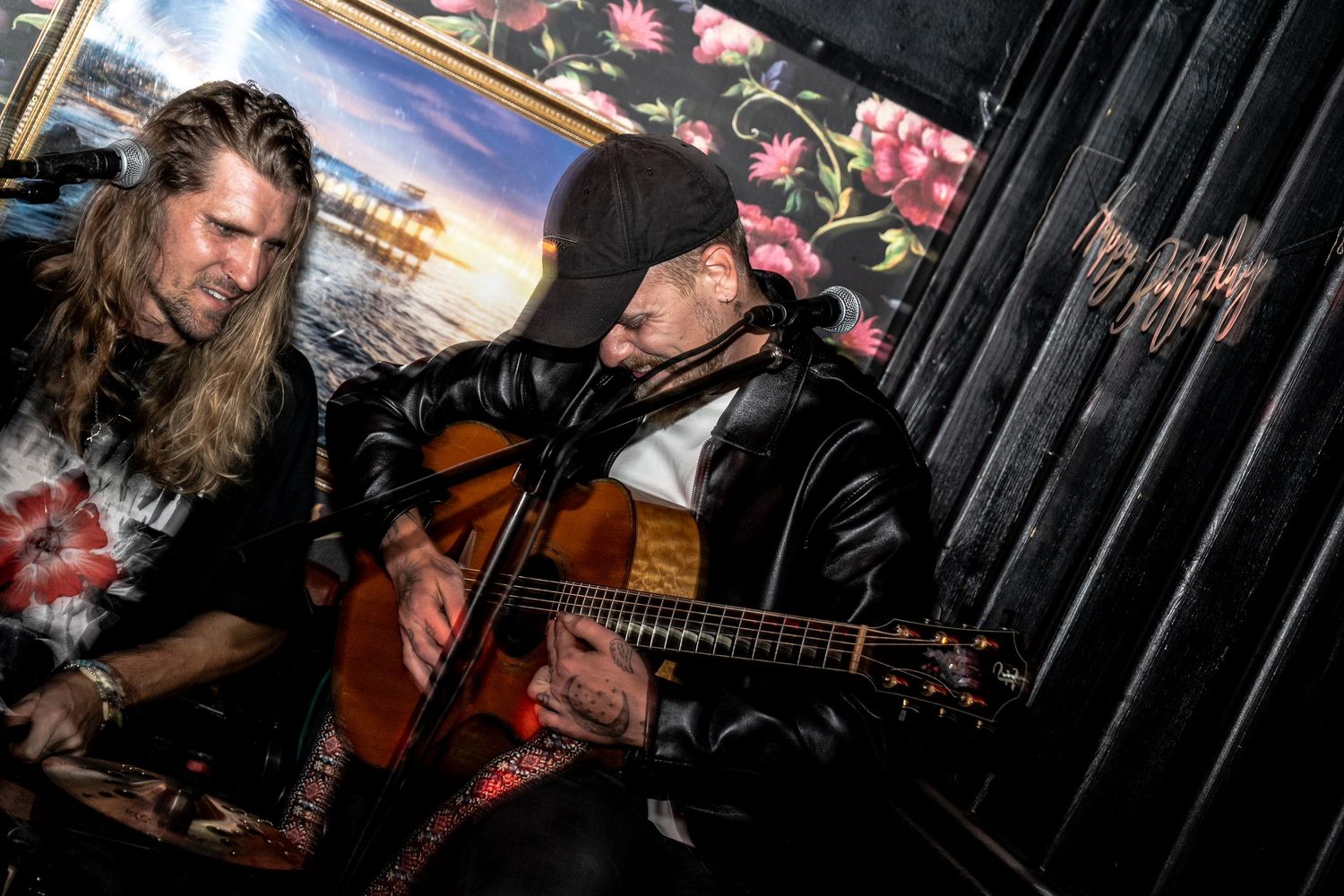 Two musicians perform on stage, one with long hair and one in a cap and leather jacket playing acoustic guitar by a microphone.