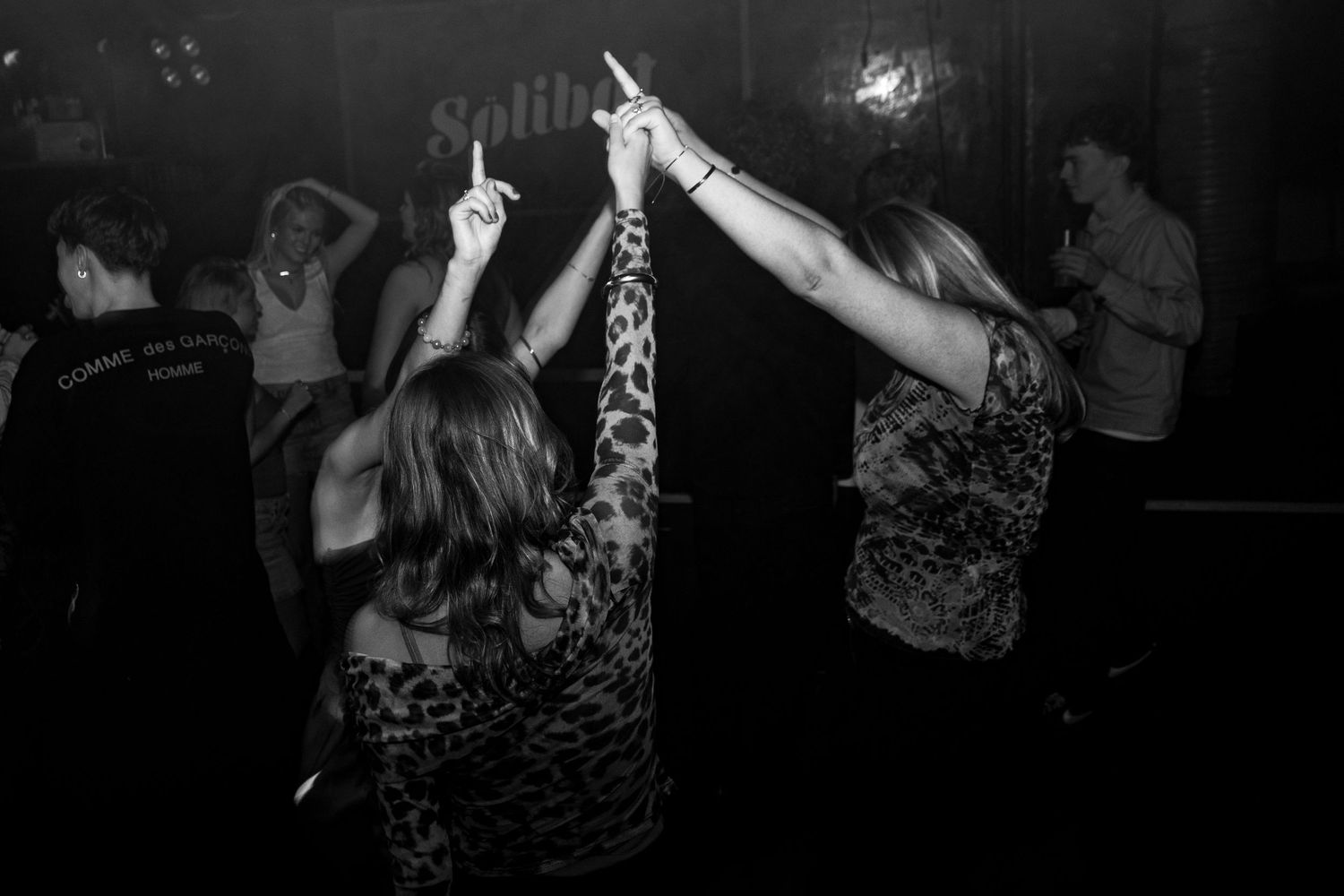 Black and white photo of two women raising their arms while dancing in a dark nightclub with people in the background.
