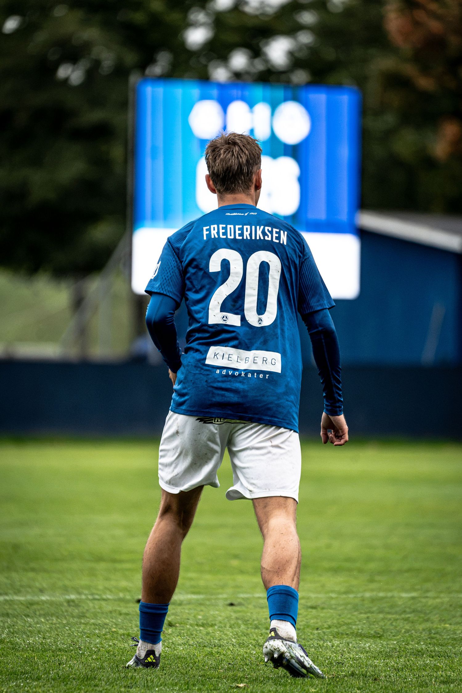 Soccer player Frederiksen number 20 in blue kit walks on green pitch during a match.
