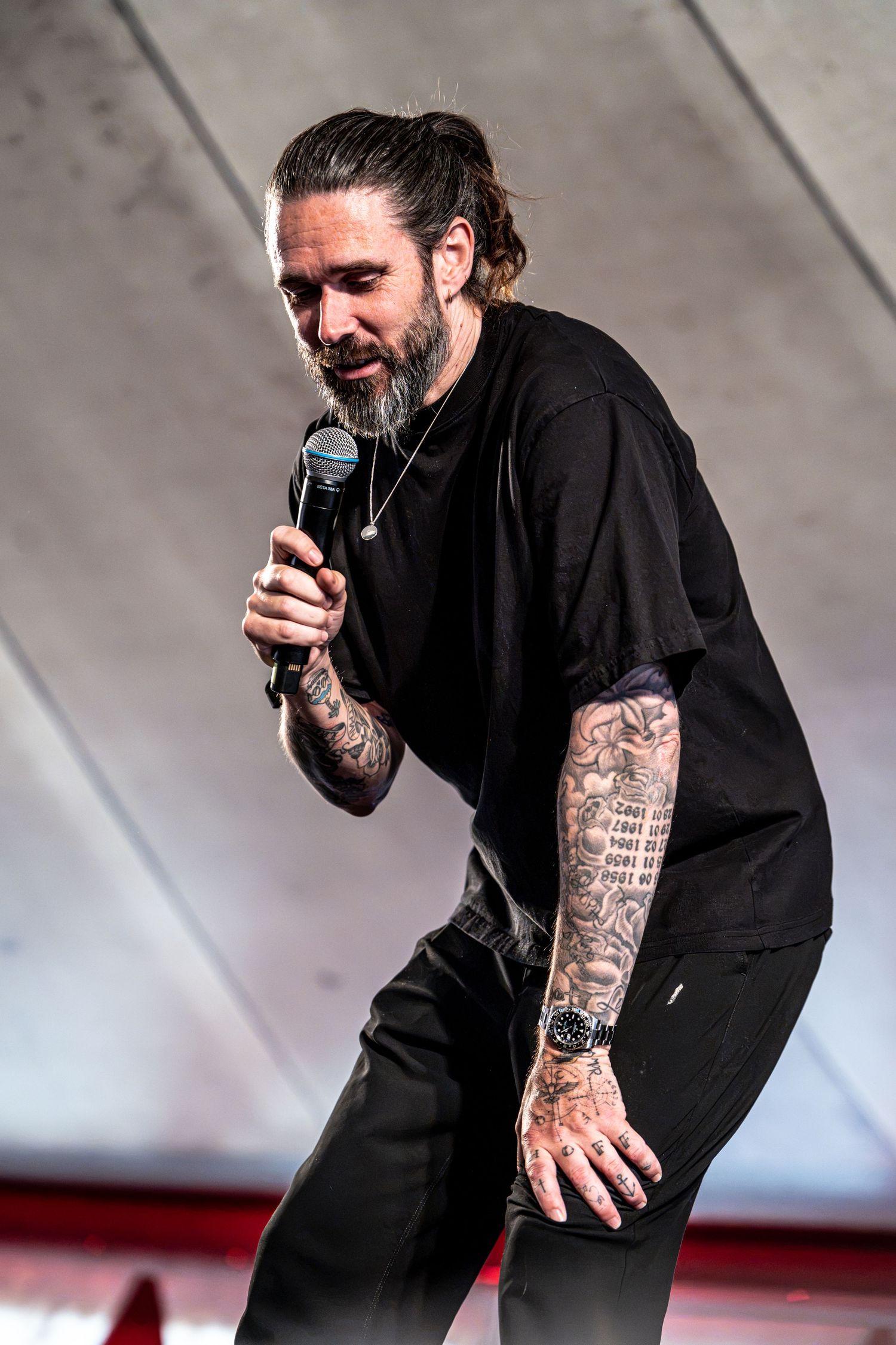 Bearded tattooed male performer bowing forward while holding microphone on outdoor stage.