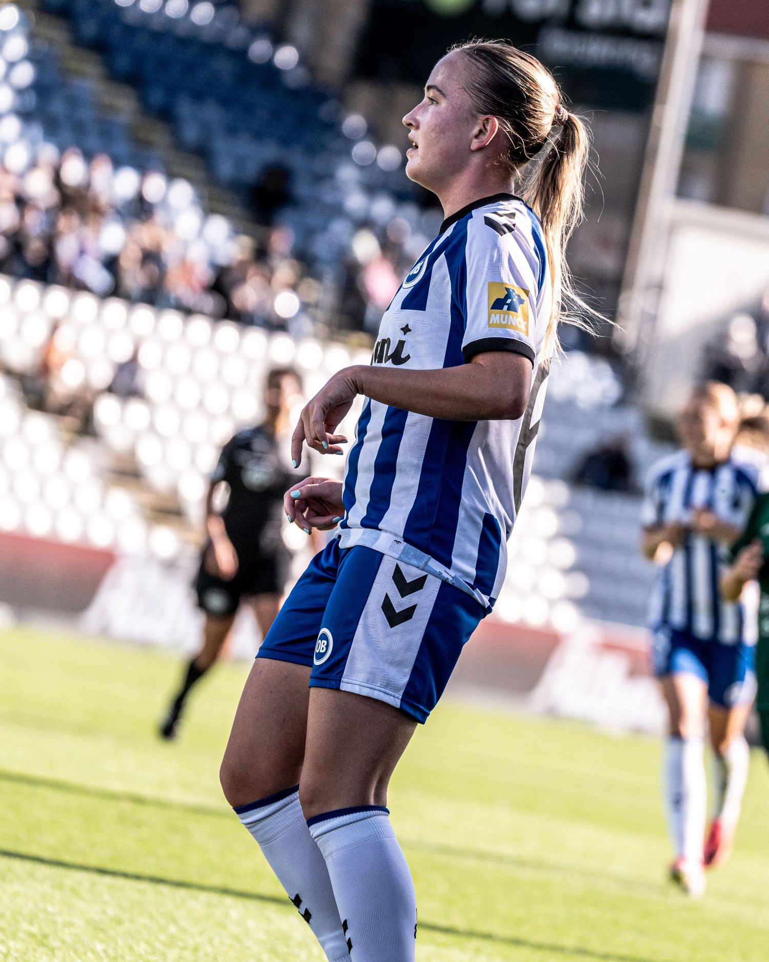 Female soccer player in Hummel blue and white kit with ponytail looking up during a sunny stadium match.