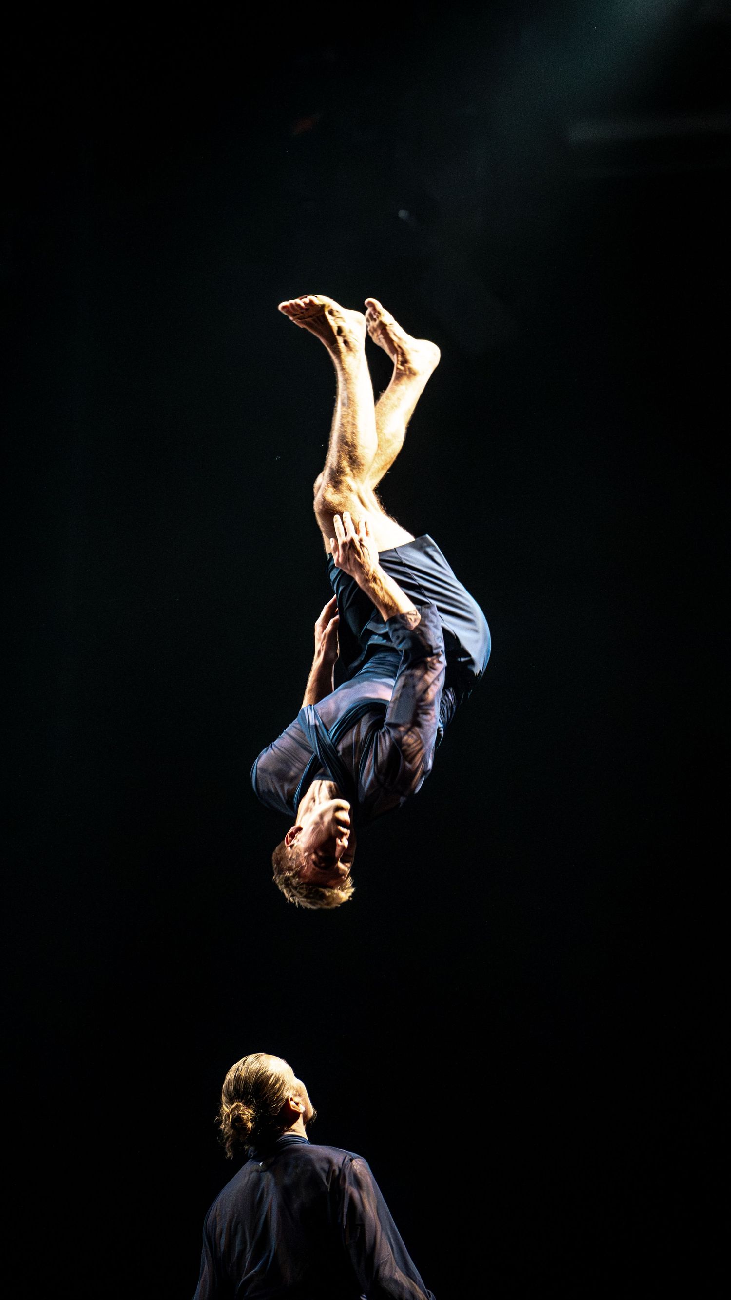 Male acrobat flipping upside down high above a performer on a dark theatrical stage.