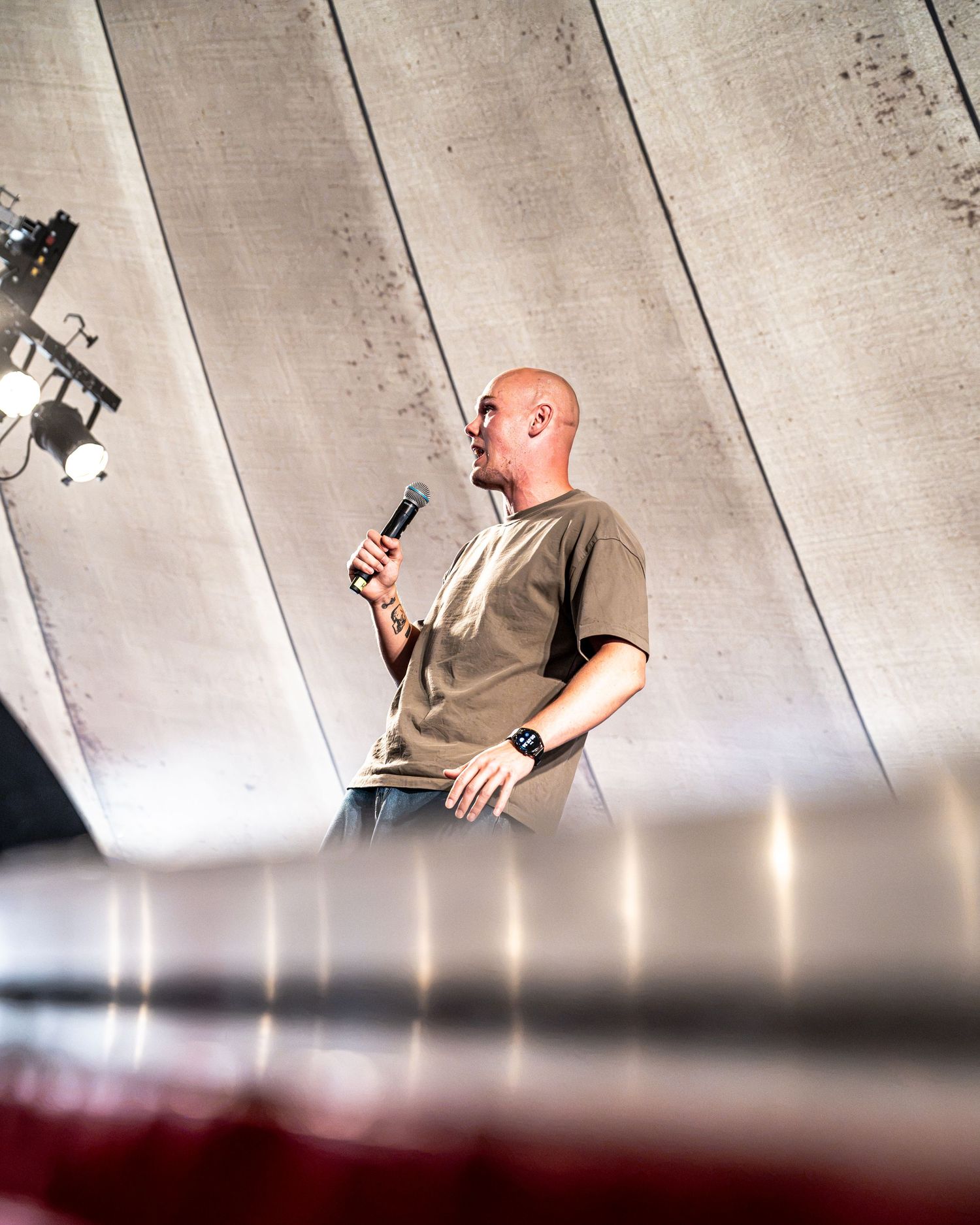 Bald male performer holding a microphone on stage with dramatic lighting.