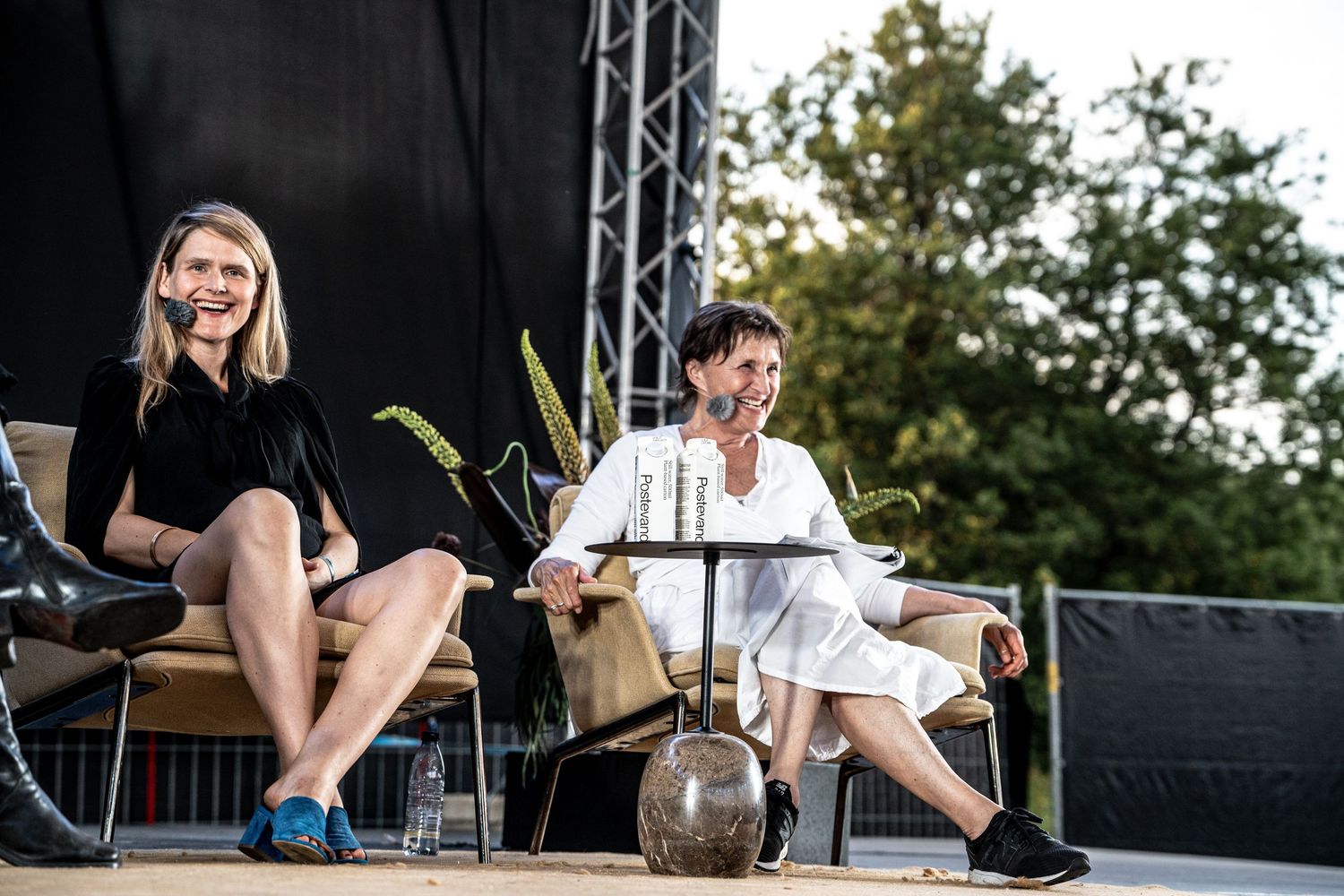 Two women sitting on chairs on an outdoor stage smiling during a panel discussion at a summer event.