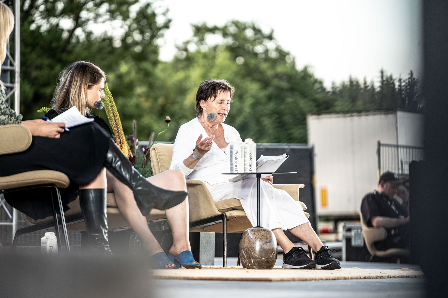 Two women in conversation on an outdoor stage, one in white dress gesturing expressively.