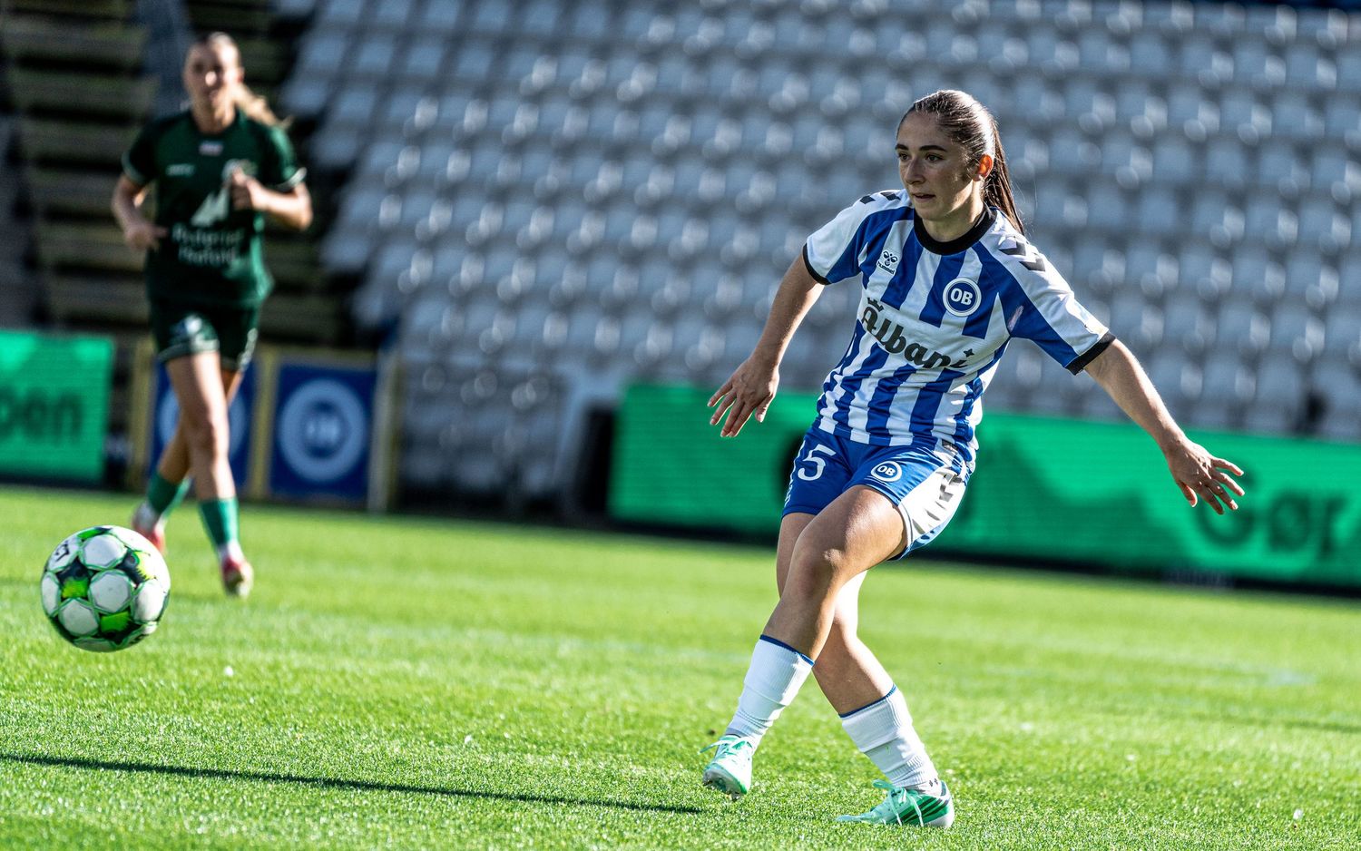 Female soccer player wearing number 5 blue and white jersey passes ball during sunny stadium match.