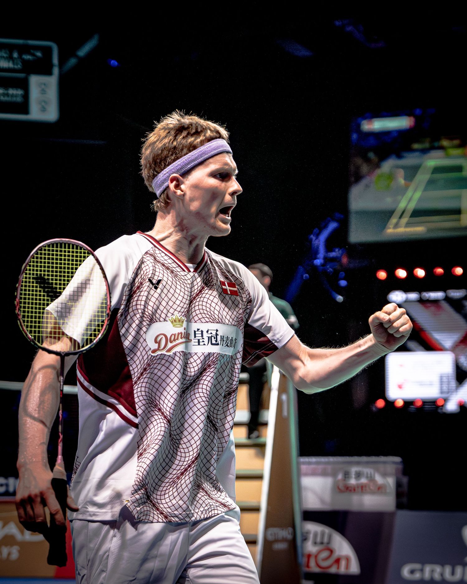 Anders Antonsen celebrating victory with fist pump wearing Denmark national badminton team jersey under arena lights.