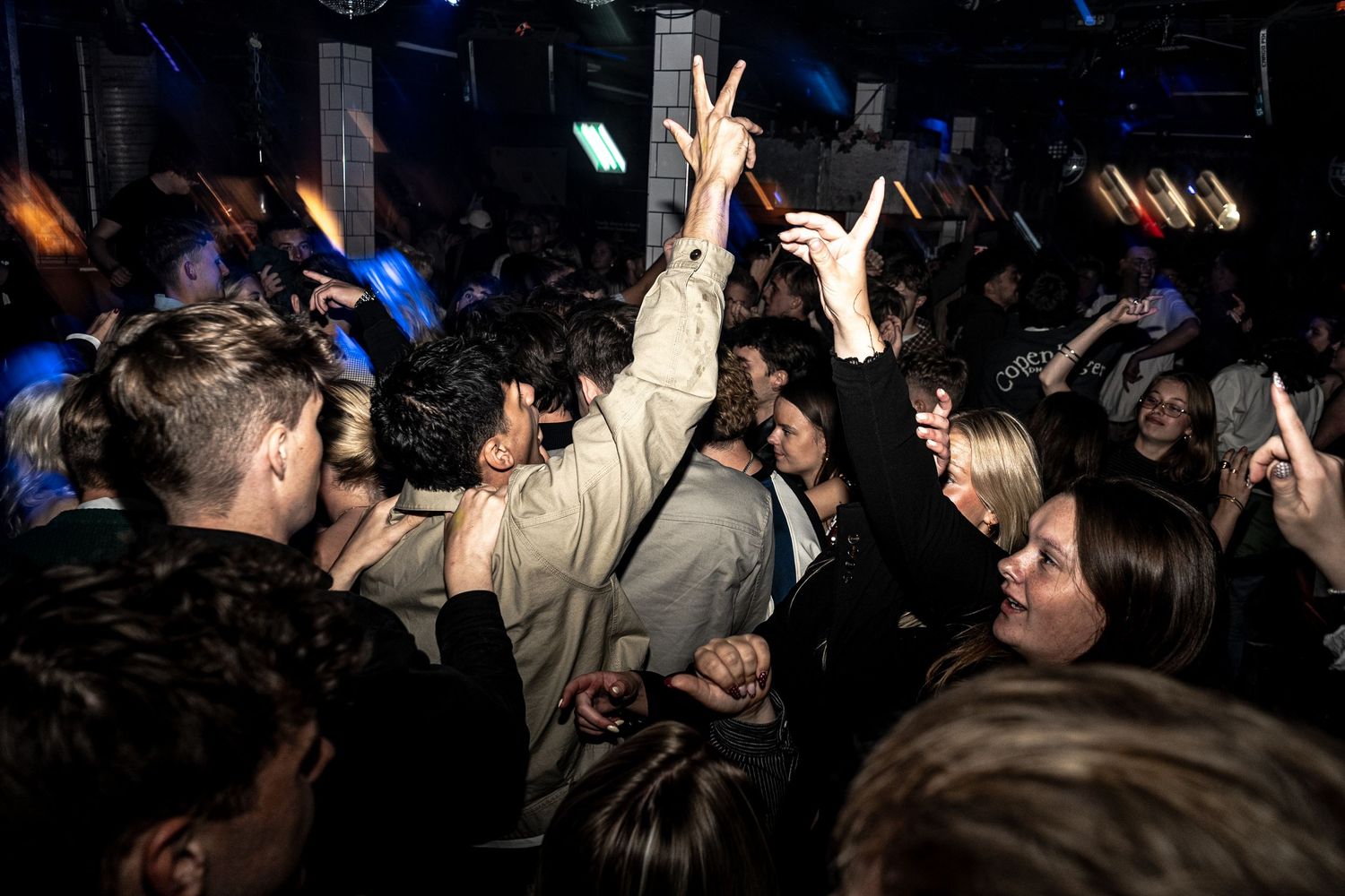Crowded nightclub dance floor with partygoers raising their hands and dancing under colorful stage lights.