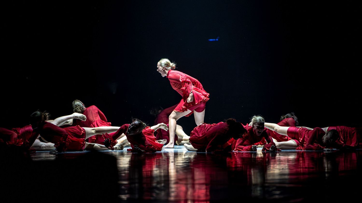 Dancers in red costumes perform a dramatic group routine on a dimly lit stage.