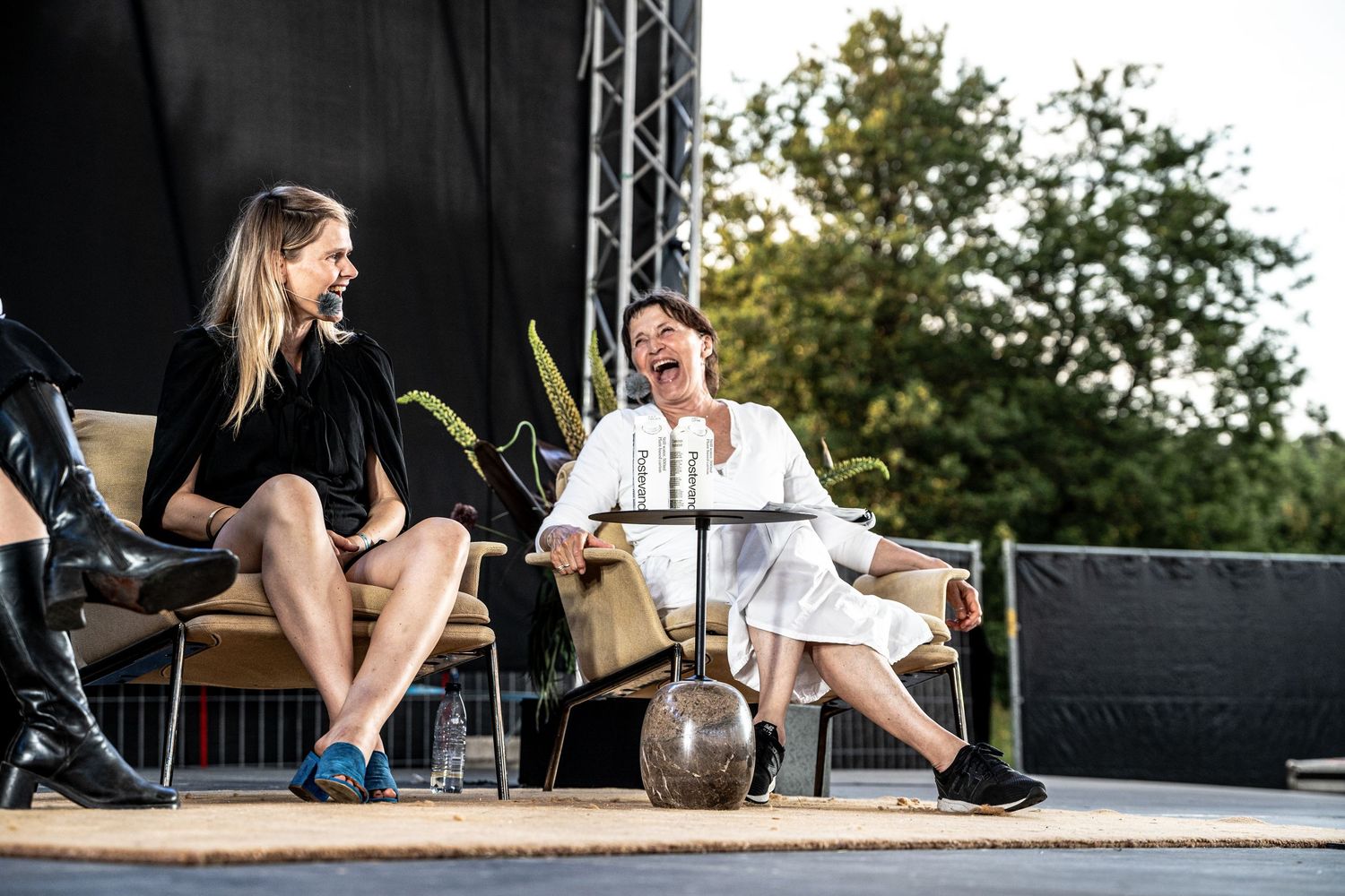 Two women smiling and conversing on stage at an outdoor panel event in natural daylight.