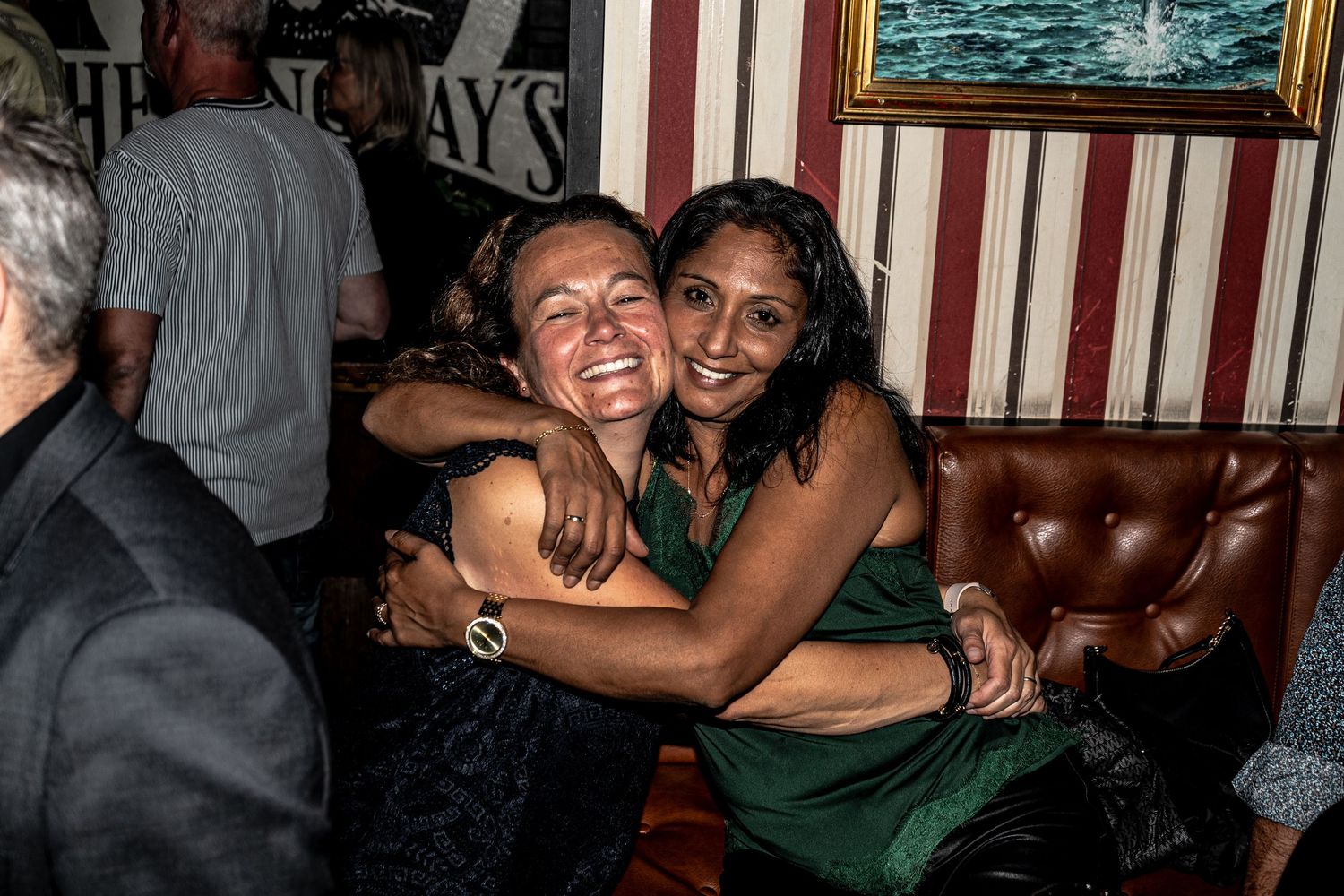 Two smiling women hug warmly on a brown leather bench in a cozy, striped-wall pub during a festive night out.