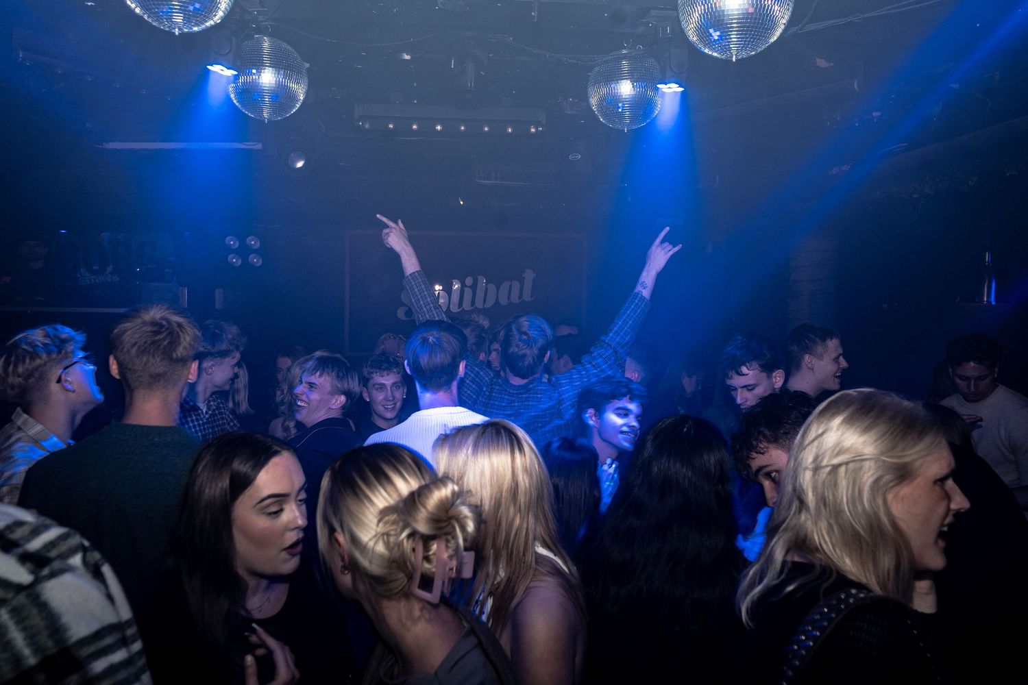 Crowd of people dancing and cheering with raised arms in a blue-lit nightclub under hanging disco mirror balls.