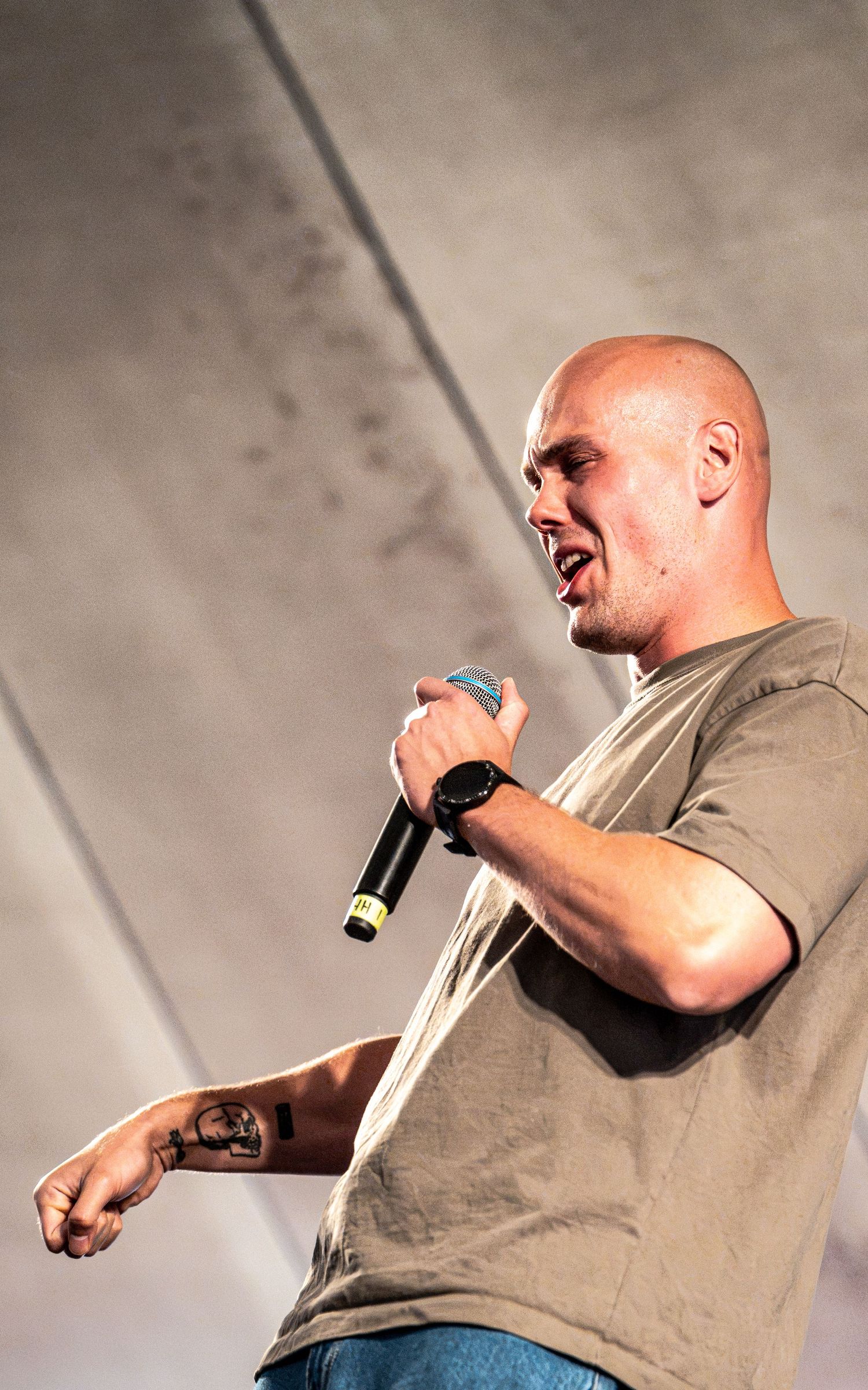 Bald male rapper with tattoos performs energetically on stage holding a microphone at an outdoor festival.