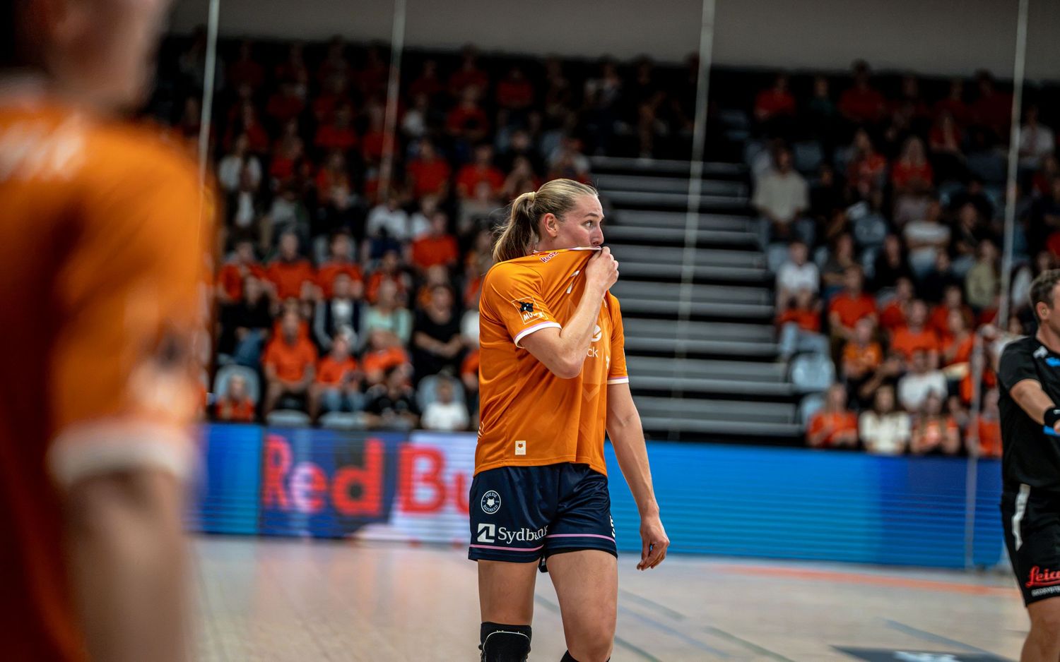 Female handball player in orange jersey wipes her face during a break in an indoor handball game.