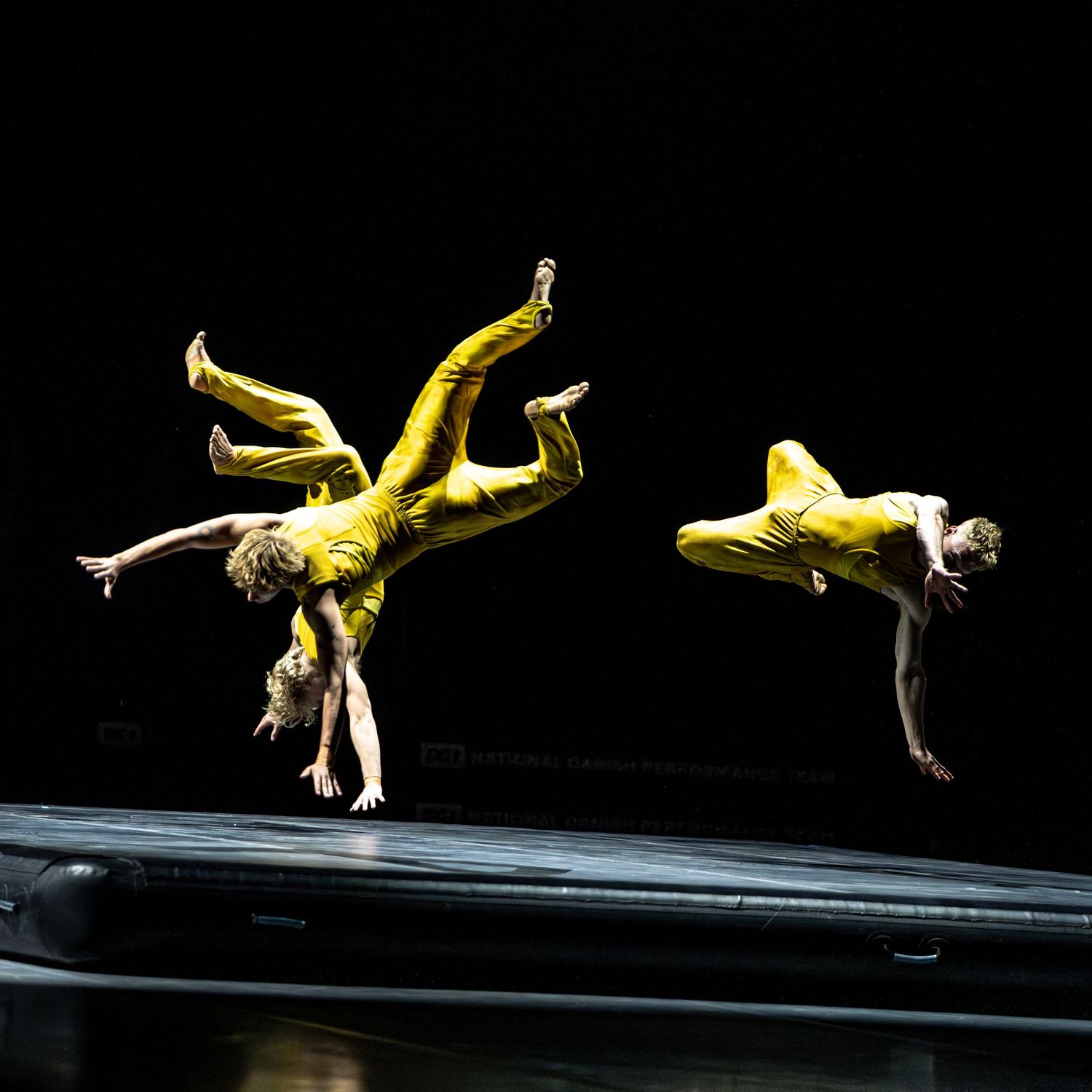 Two acrobats in yellow suits perform mid-air flips on trampoline against black background.