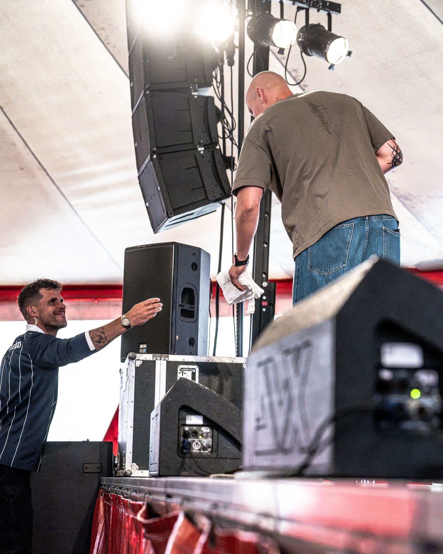 Bald male performer leans down to interact with crew member beside stage speakers at outdoor festival.