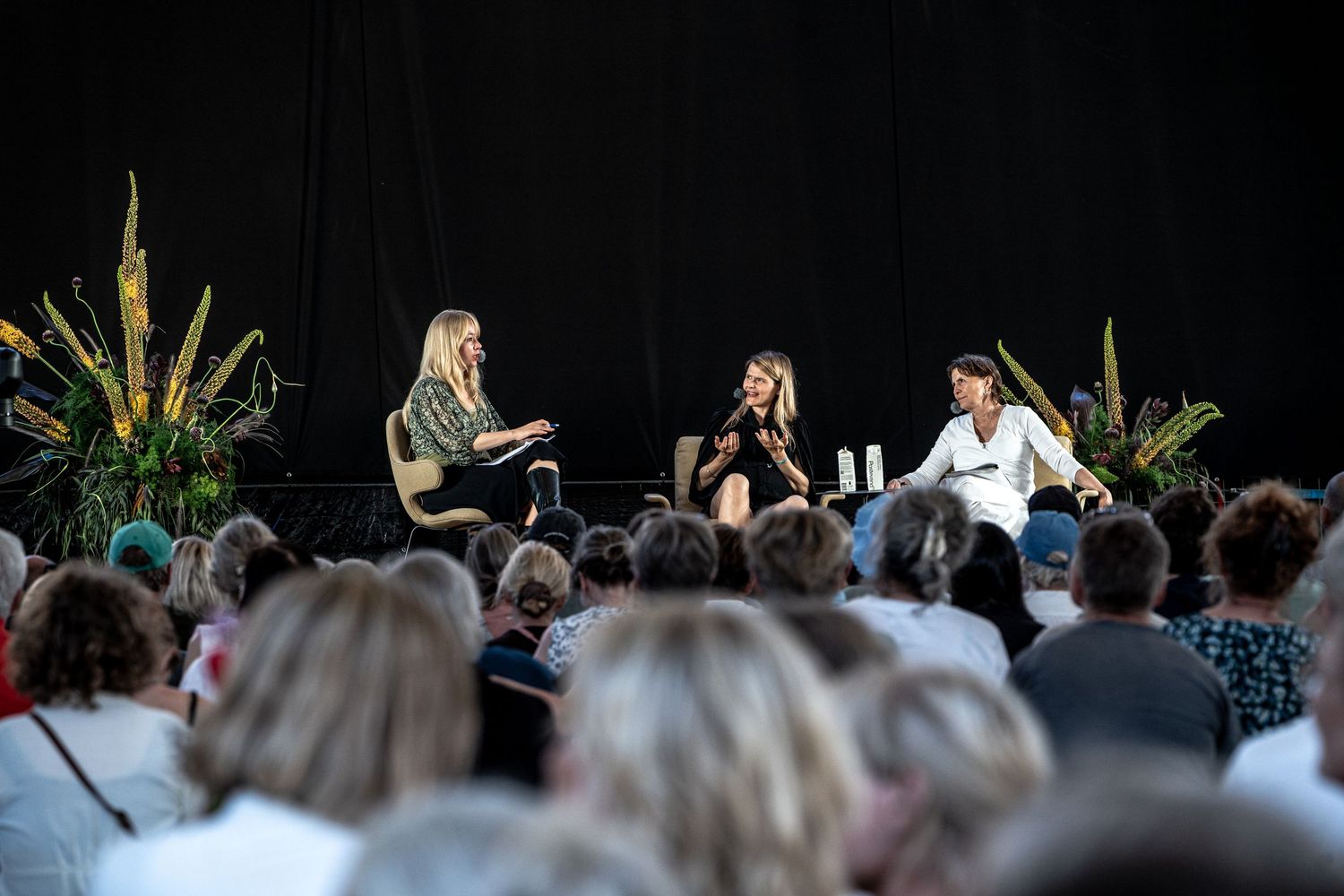 Three female panelists on stage with tropical plants at outdoor event.