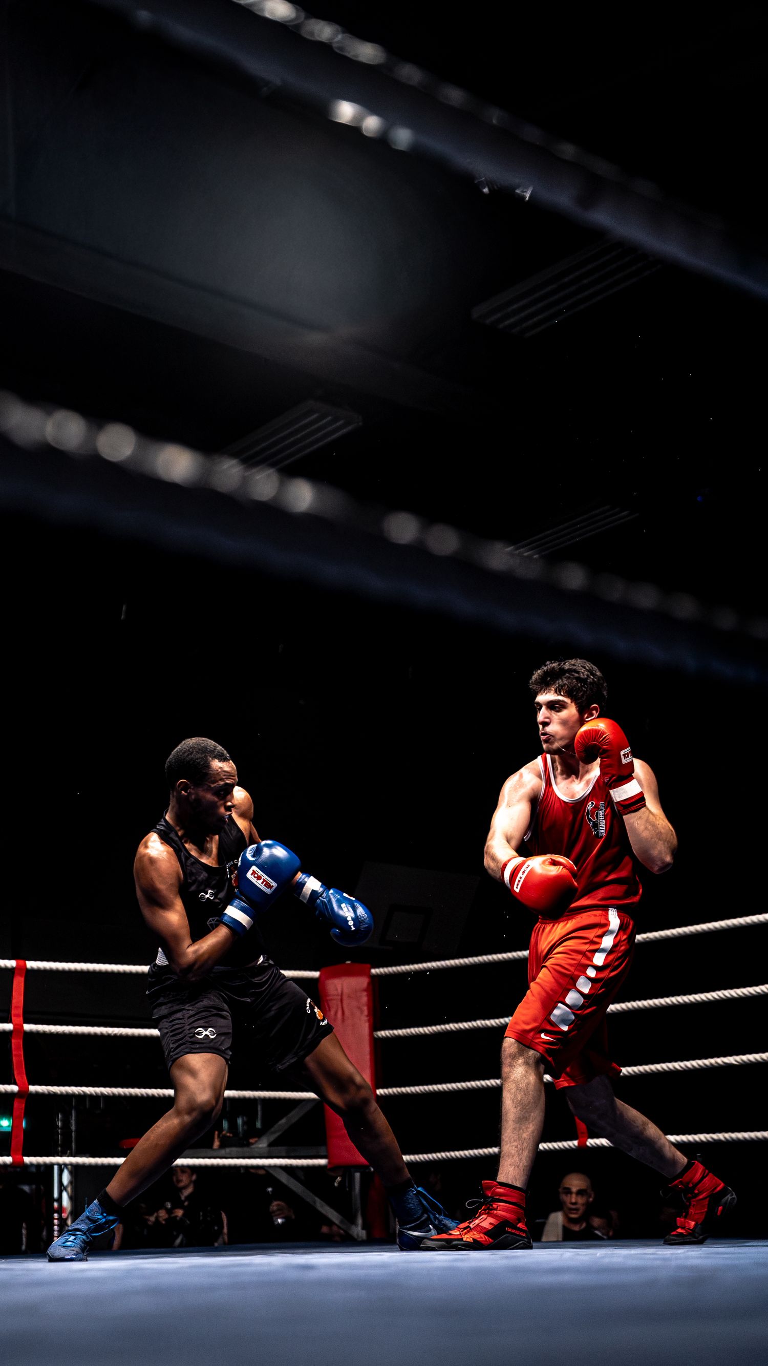Two boxers compete in a professional boxing match, one in red and one in black, exchanging punches in a dark arena.