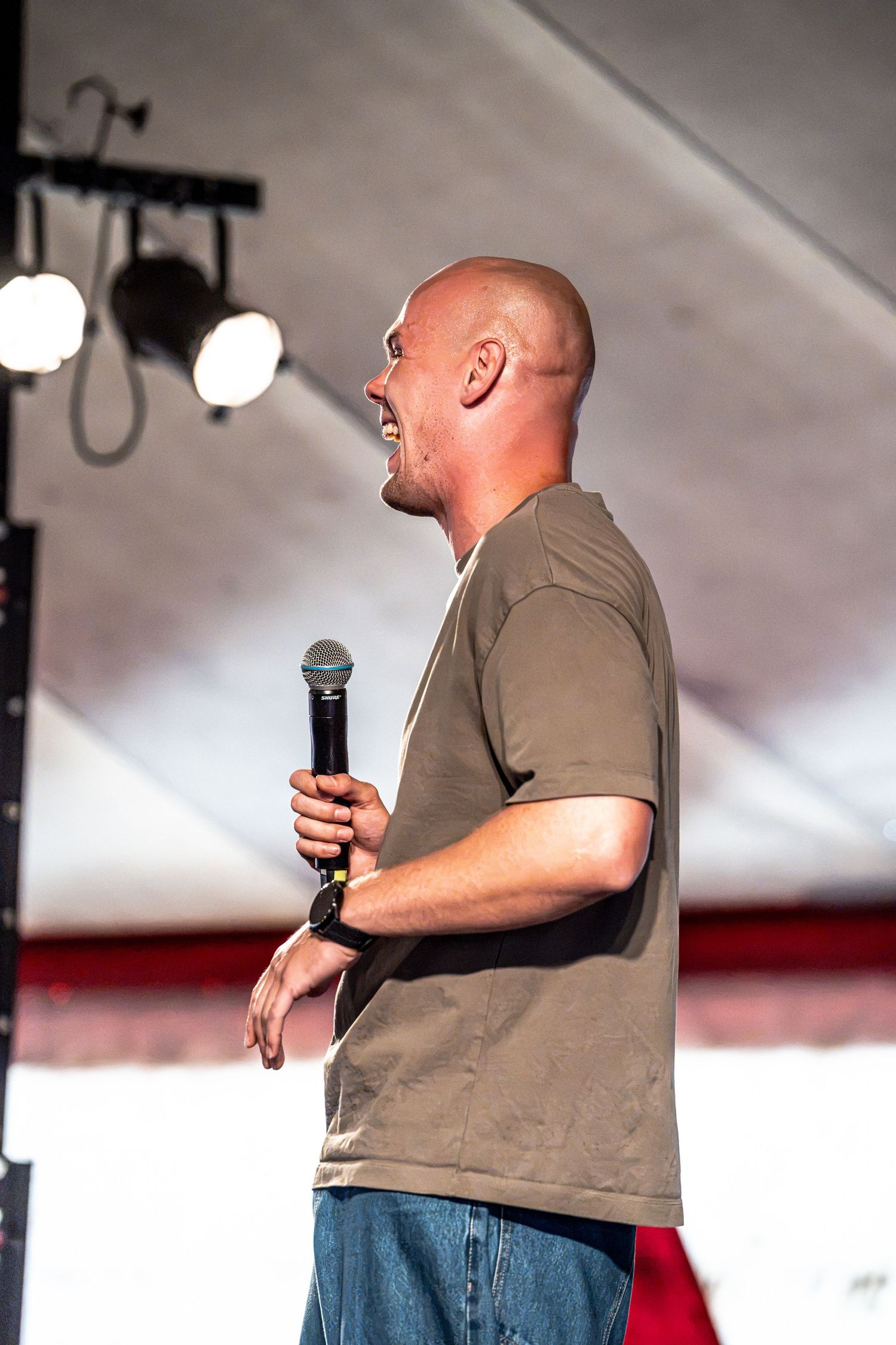 Bald male performer laughs while holding microphone on stage at an outdoor event under a white tent.
