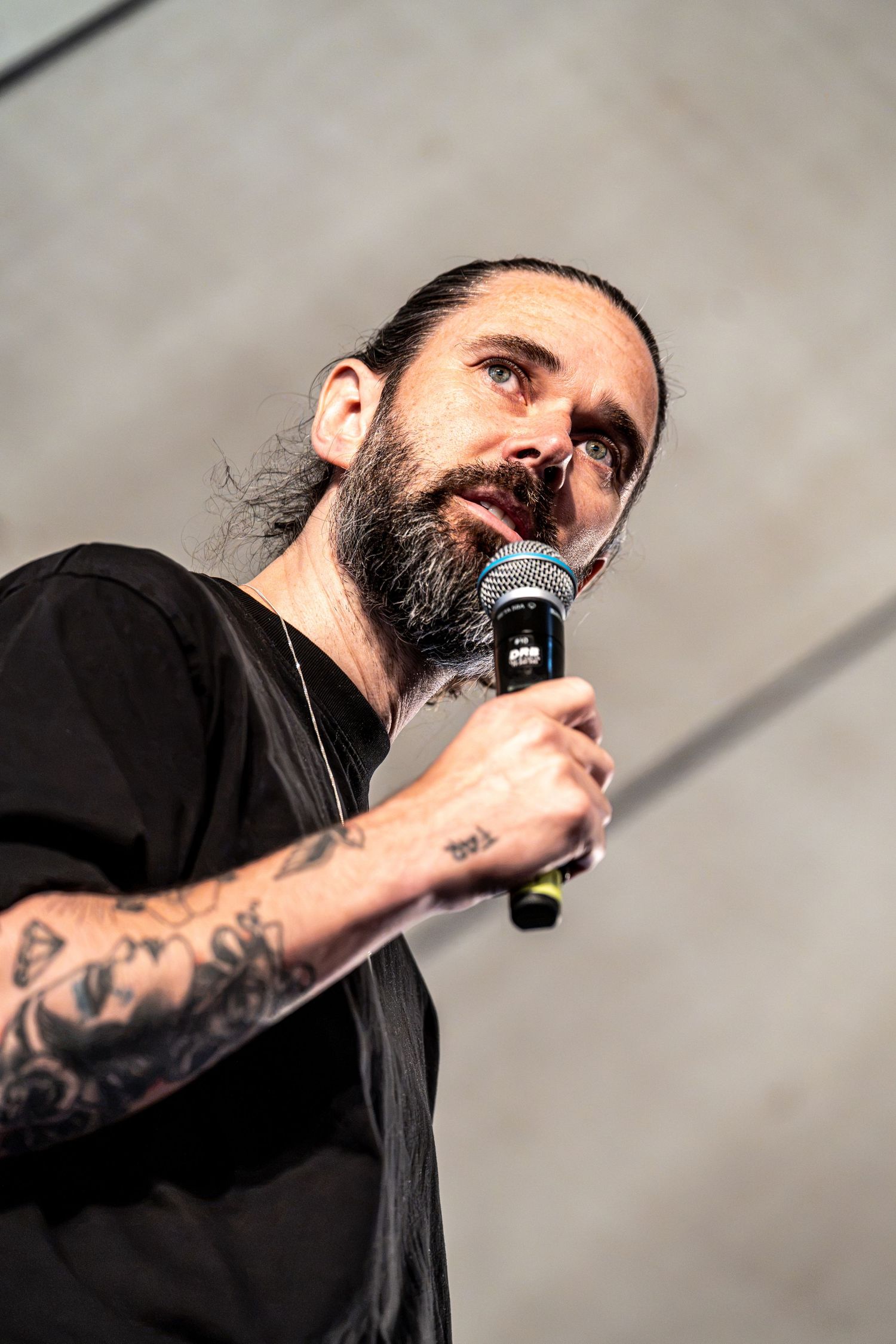 Bearded tattooed vocalist looks up while singing into microphone on outdoor stage.