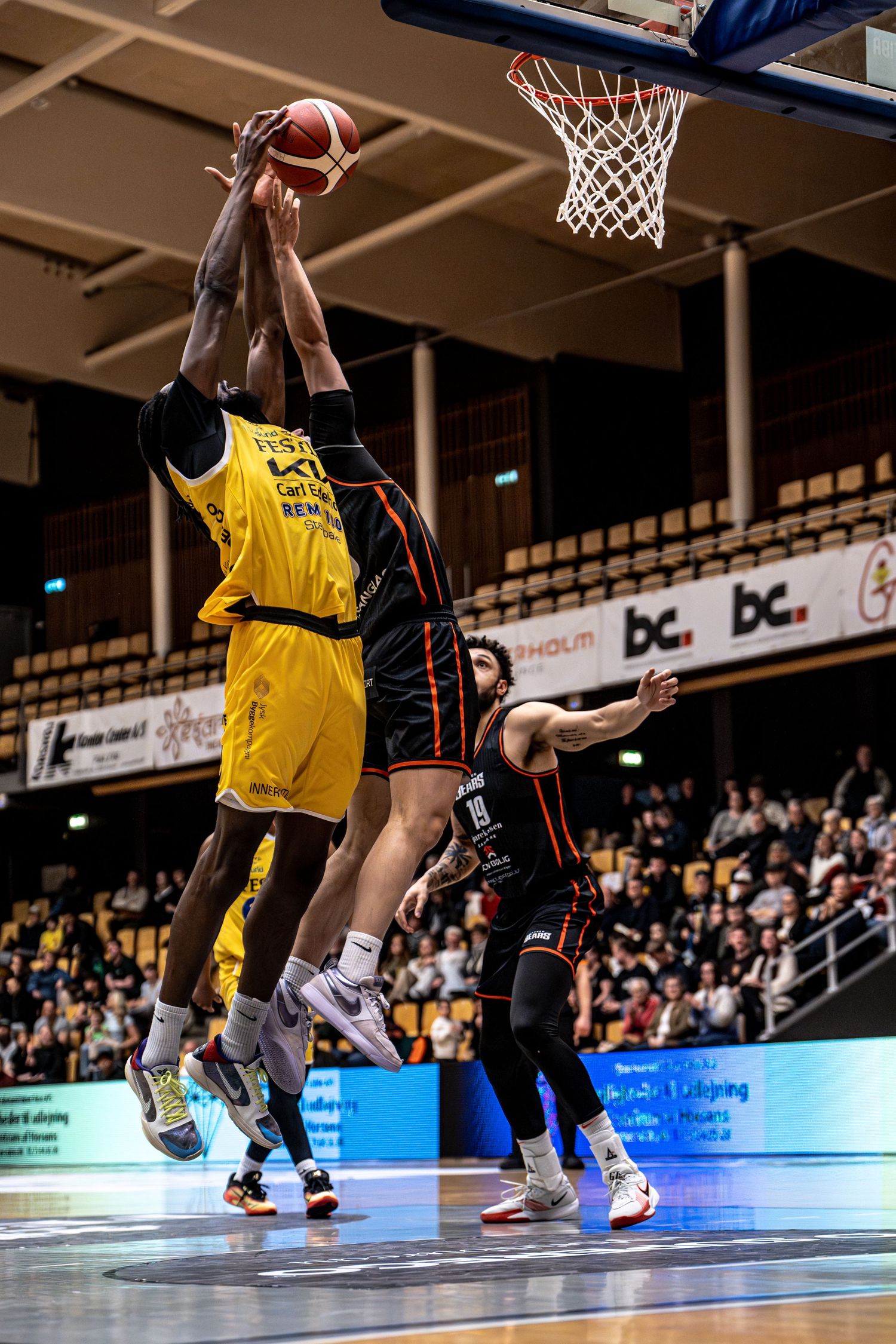 Yellow team player attempts a layup against black-uniformed defender in packed basketball arena.