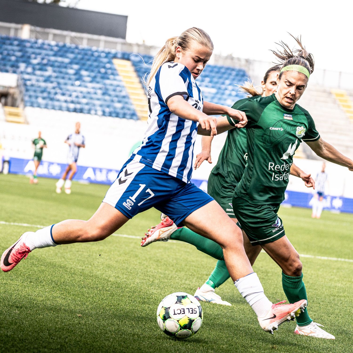 Two female footballers compete for the ball, one in blue and white, the other in green, during a match.