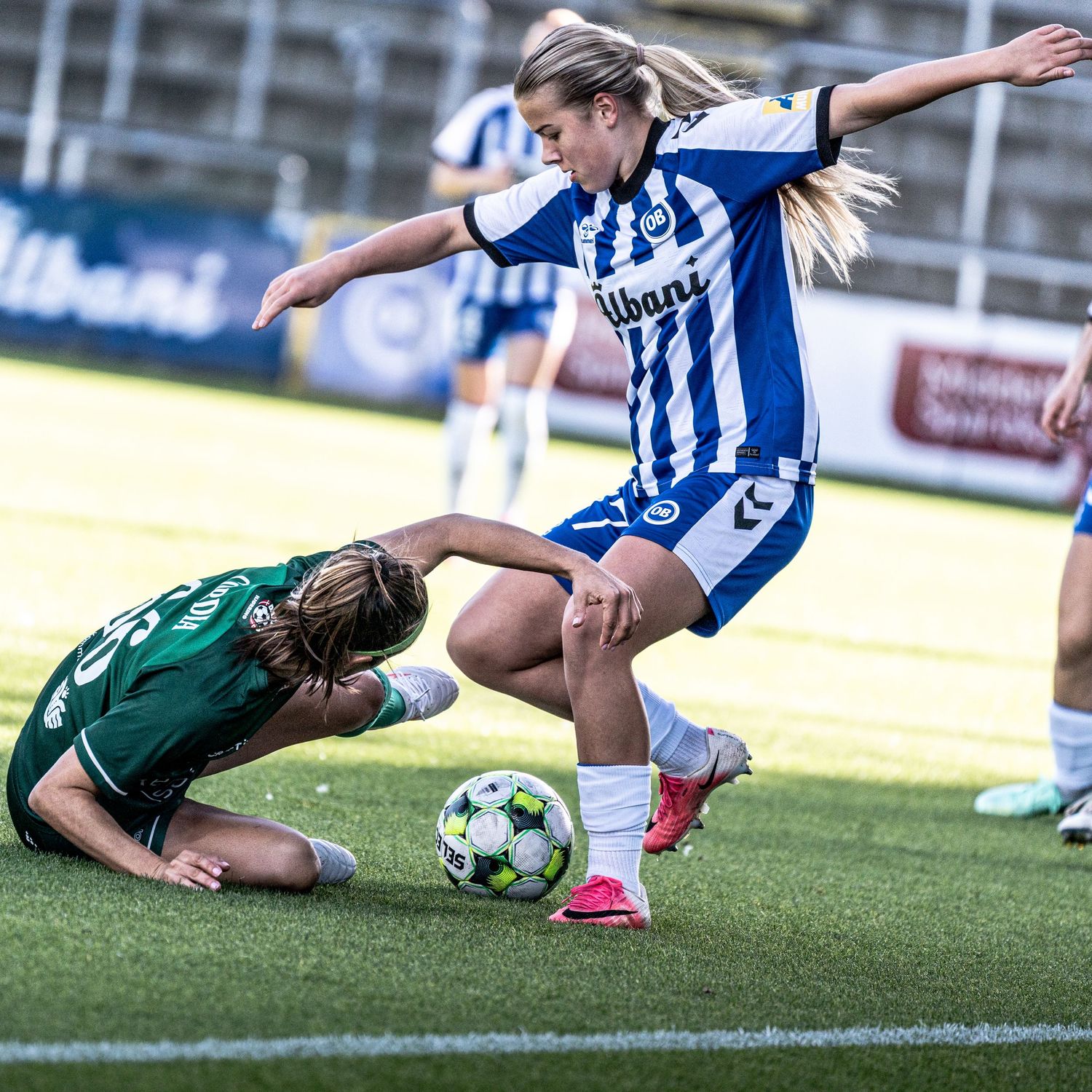 Two female soccer players competing for ball during match in blue and green kits.