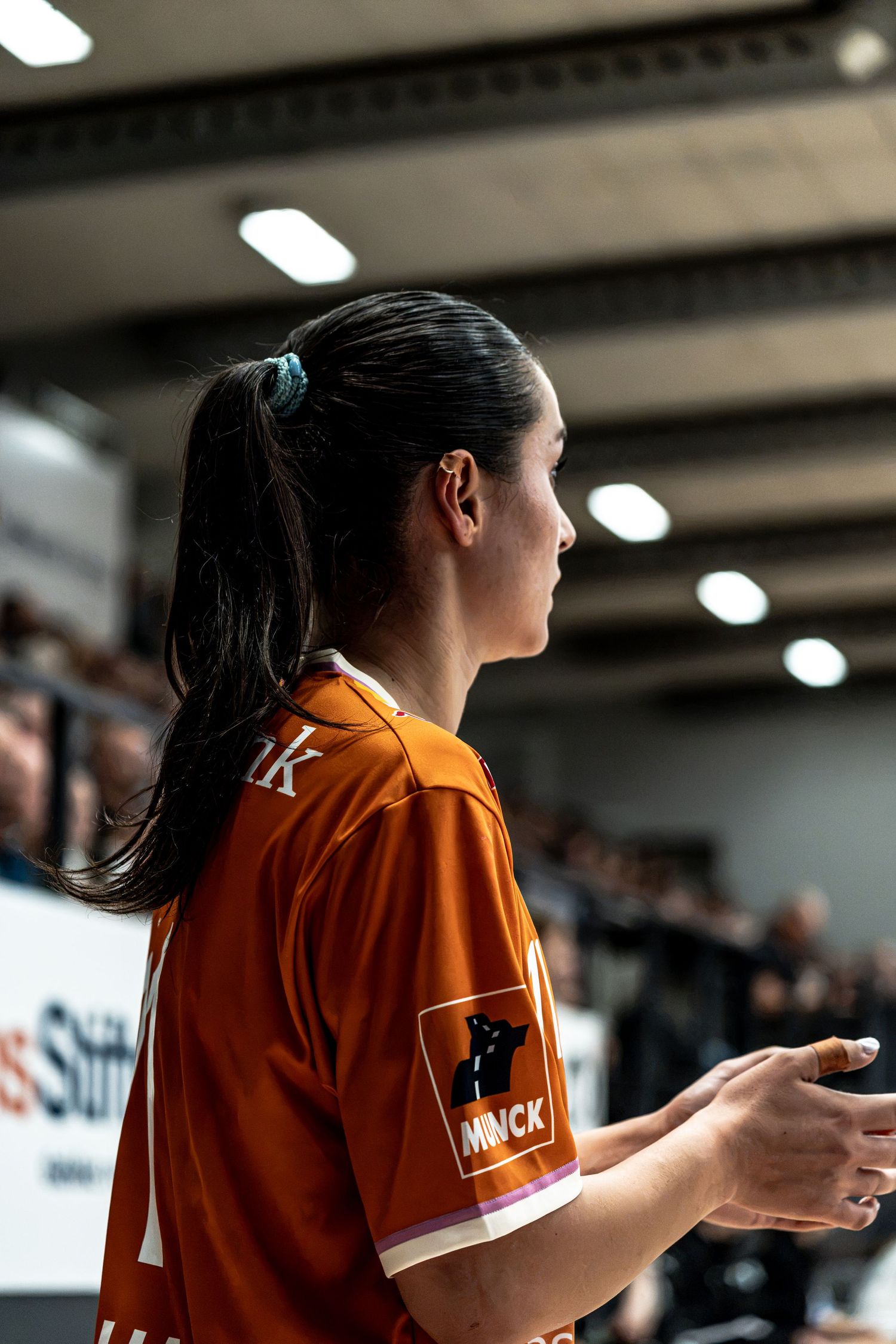 Female handball player in orange jersey with ponytail standing on the sideline during an indoor handball game.