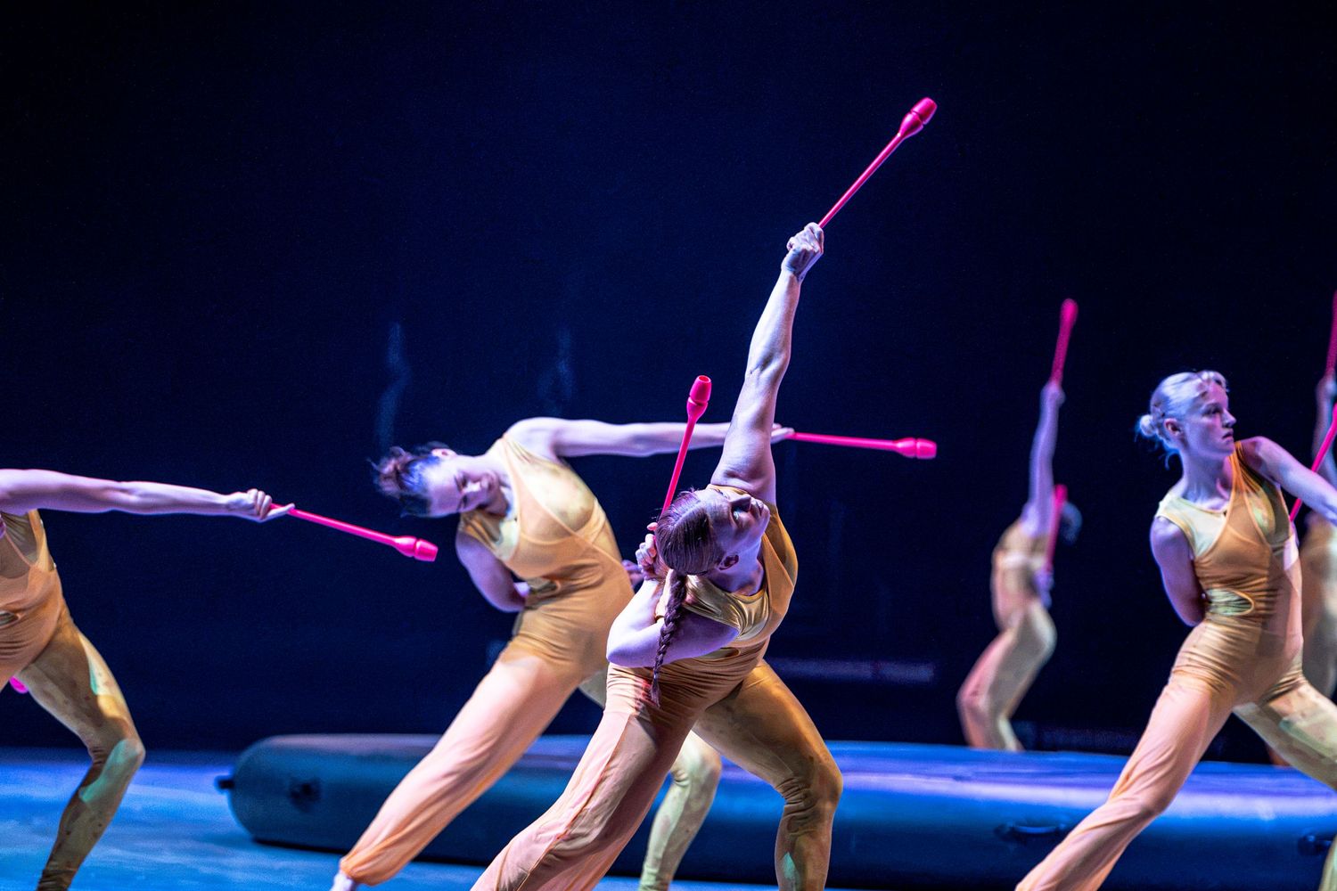Rhythmic gymnasts in nude costumes execute elegant poses with pink clubs on blue-lit stage.