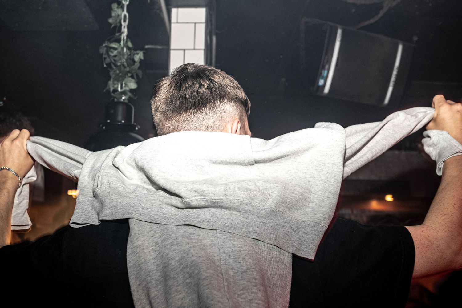 Man seen from behind pulling a gray sweatshirt over his head with raised arms in a dark nightclub with stage lighting.