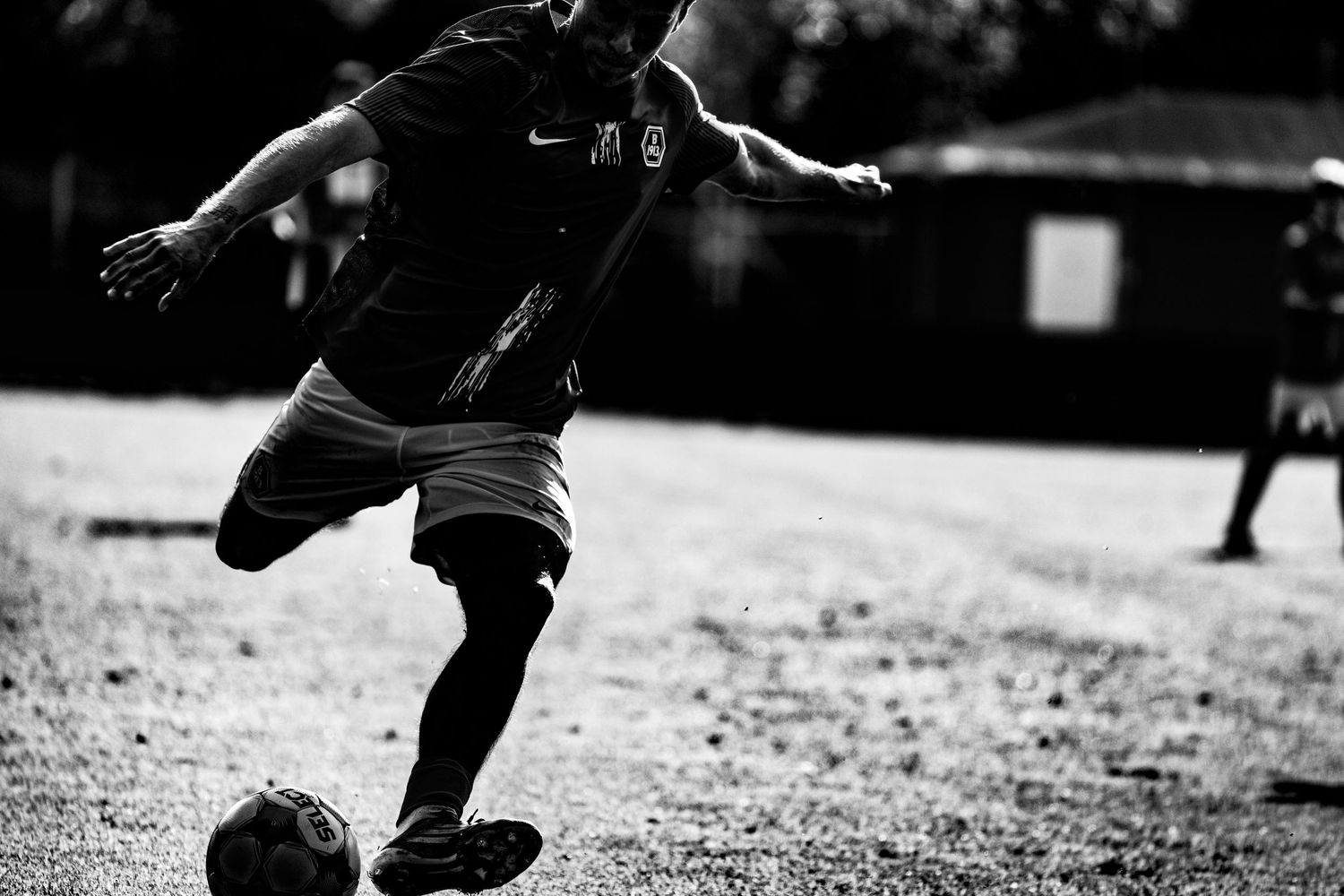 Black and white photo of soccer player kicking ball on dirt field during outdoor match.