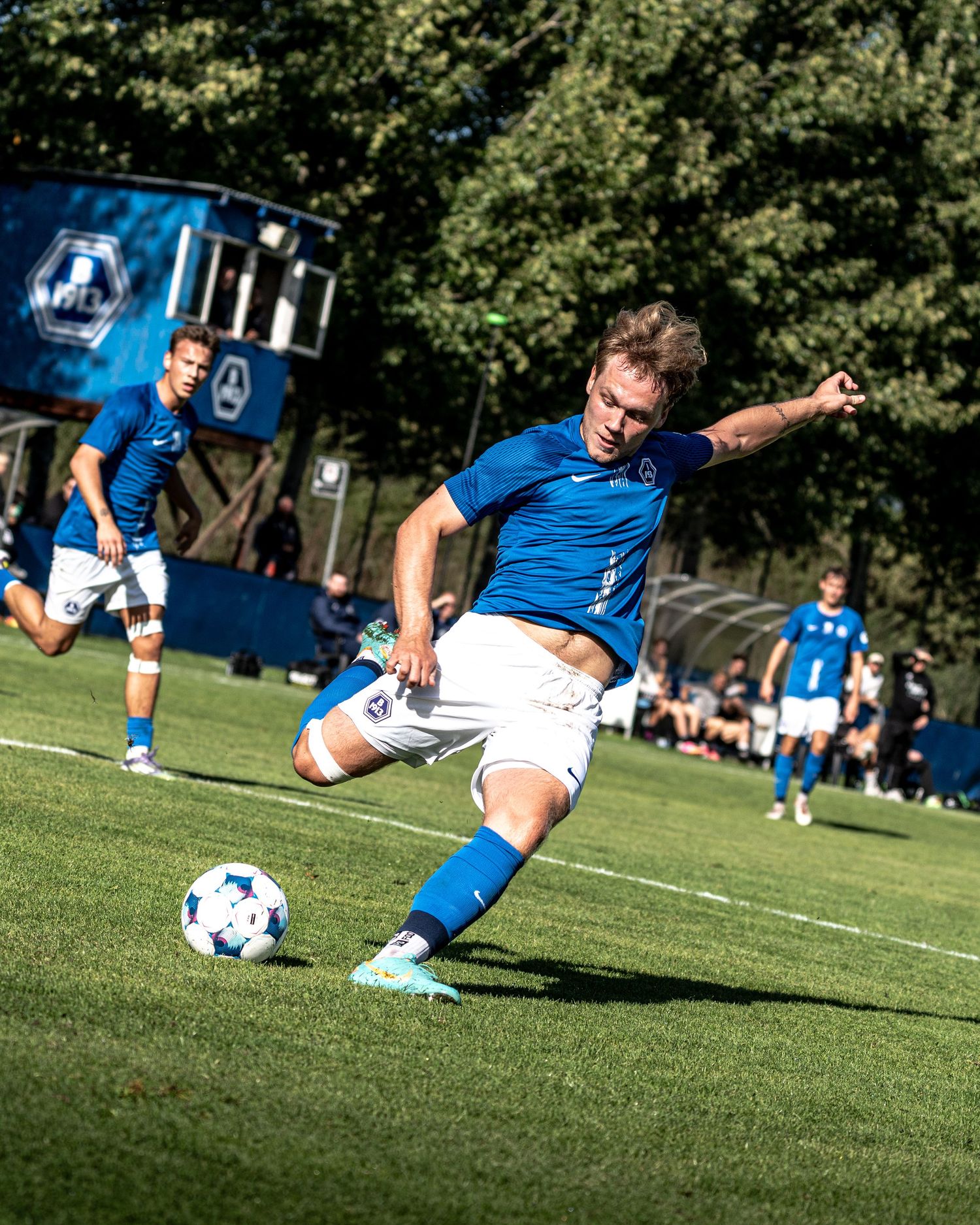 Soccer player in blue Nike kit striking ball on green field during sunny outdoor game.