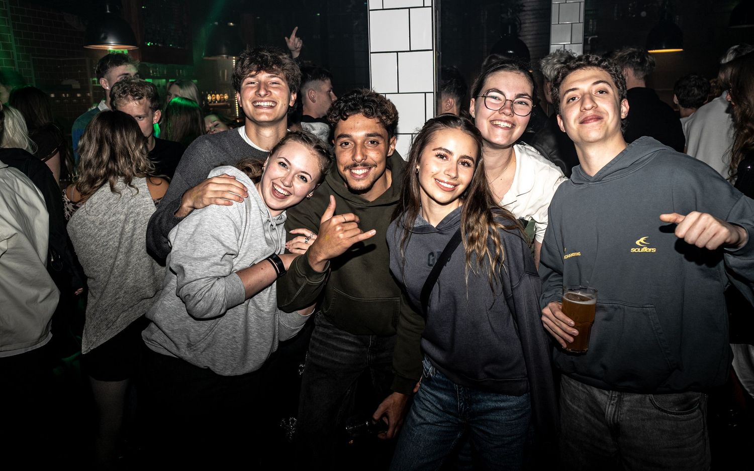 A cheerful group of friends huddles together at a lively bar, smiling and gesturing at the camera while one man holds a beer.
